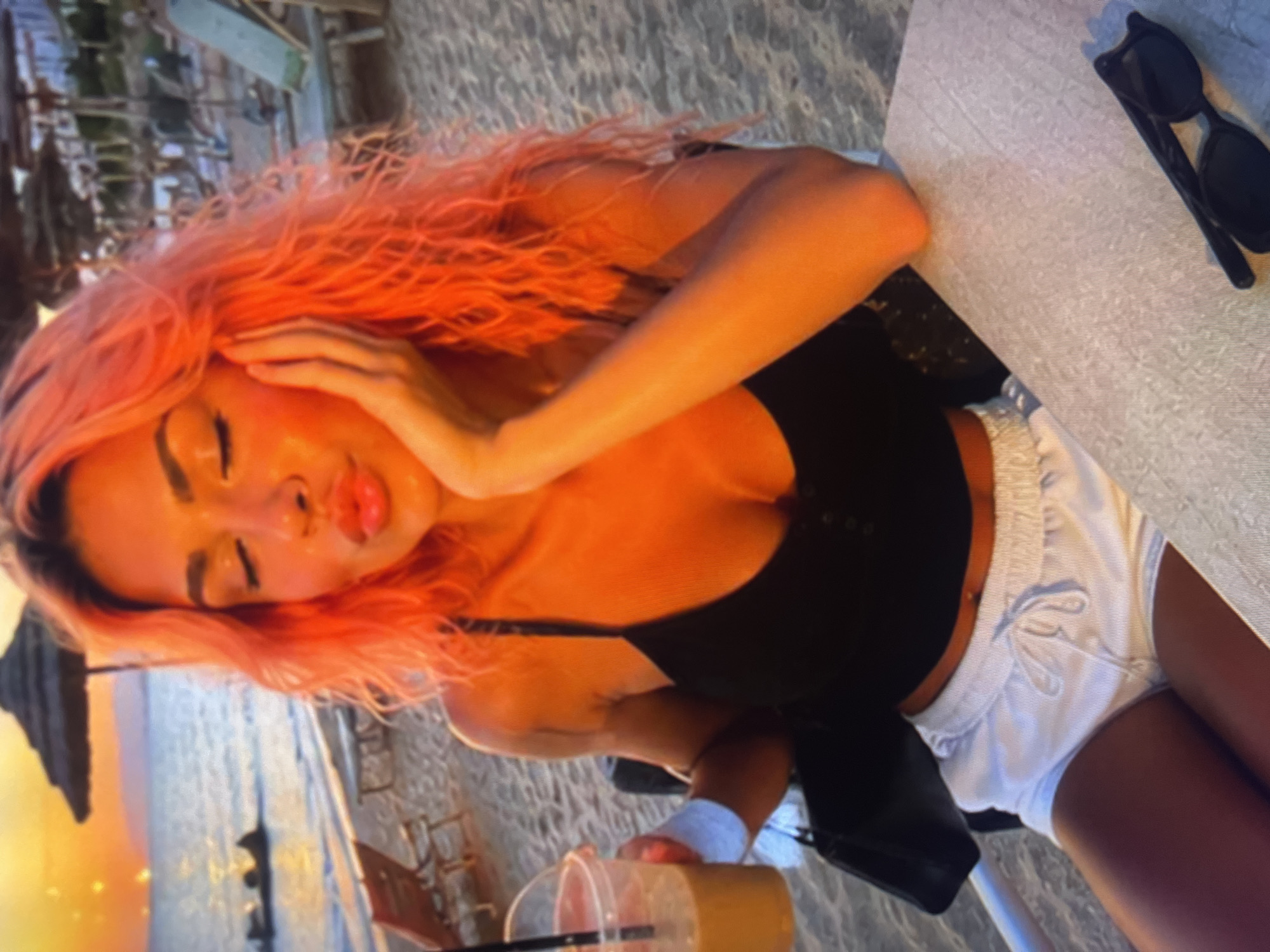 In the city of Unknown, Unknown, at a beachside location during sunset, a young woman with vibrant pink hair sits at a table on the sand. She is wearing a black tank top and white shorts, and her hand is resting on her cheek as she puckers her lips. A plastic cup with a beverage and straw is held in her other hand, and a black purse is visible beside her. Sunglasses are placed on the table in front of her. The background depicts a serene beach scene with lounge chairs, umbrellas, and the ocean reflecting the warm hues of the setting sun.