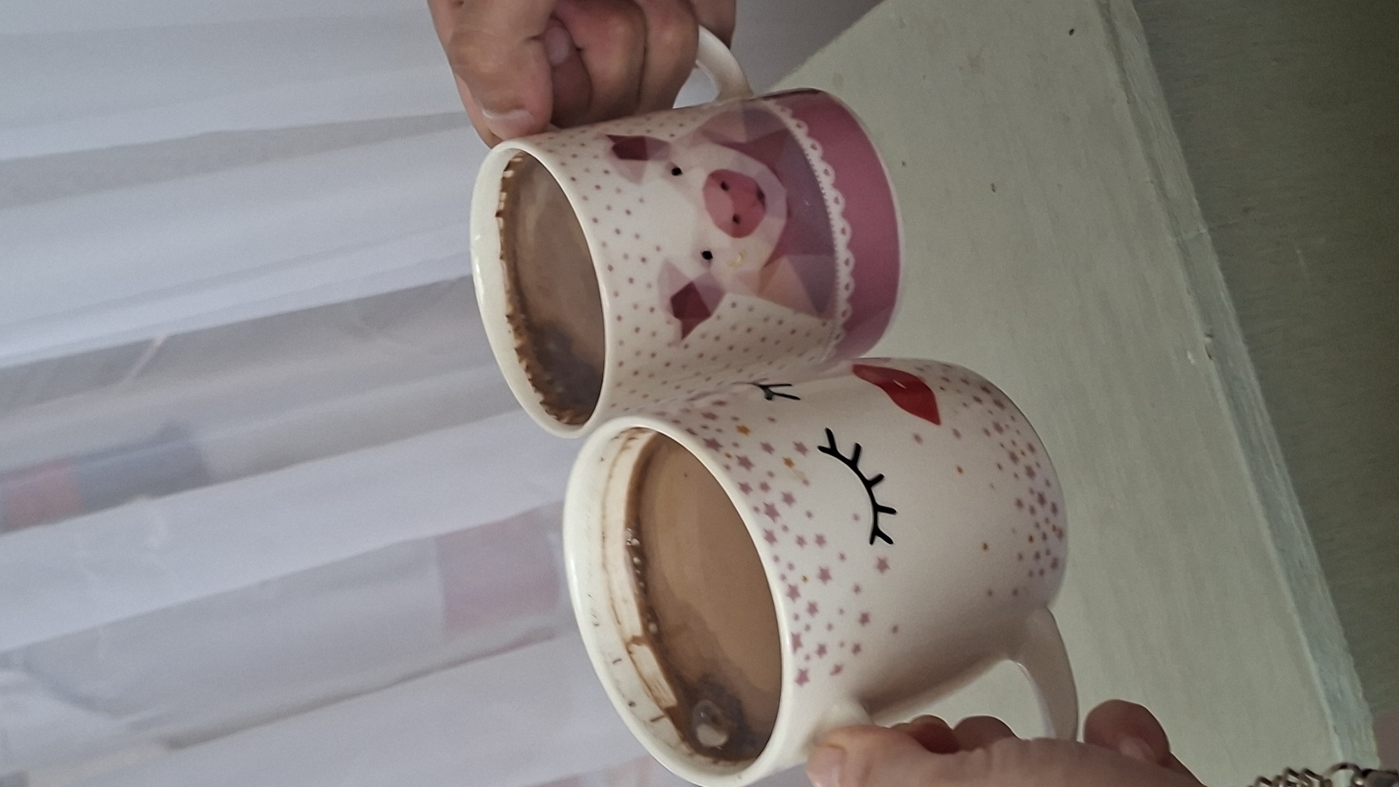 Two mugs filled with a brown liquid, likely coffee or hot chocolate, are being held up. One mug on the left features a face with closed eyes and red lips, decorated with pink stars. The mug on the right is adorned with a graphic of a pig and pink polka dots. Both mugs are white with pink accents. The background consists of sheer white curtains. The scene appears to be indoors, possibly on a shelf or table, given the visible surface in the lower portion of the image. There is no discernible information about the specific location within the city of Myadzyel, Belarus.