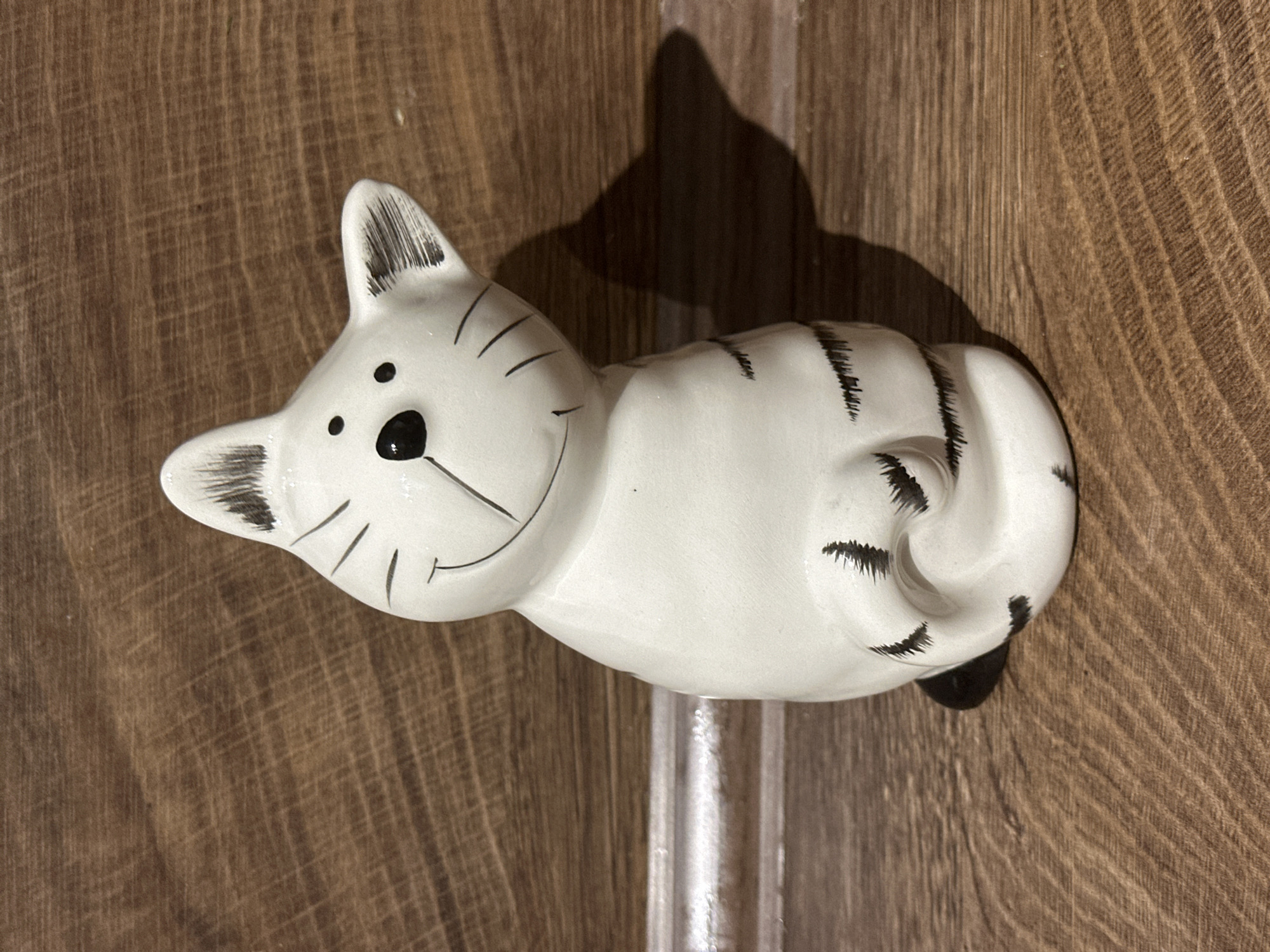 The image displays a white ceramic cat figurine with black painted details resting on a textured wooden surface. The figurine, which is the primary subject, is depicted in a relaxed, lying-down pose, curled slightly. Its face features two black dot eyes, a triangular black nose, painted whiskers, and an upturned line forming a cheerful "smile." Black stripes adorn its back and tail, and the inner ears are also detailed with black. Black paw pads are visible beneath its curled body.
The cat casts a distinct, dark shadow behind it, indicating a light source, likely from above and to the right. Beneath the figurine, a narrow, vertical, clear and reflective strip, possibly made of plastic or glass, is visible. The background is a consistent dark brown wooden surface with a prominent grain pattern running diagonally across the frame, suggesting it could be a table or shelf.
No activity or event is taking place; the image is a still life. There is no visible text in the photograph. The notable details include the figurine's stylized and smiling expression, the clear shadow it casts, and the reflective element supporting or near it. The overall setting is indoors, with the image captured in Bobov Dol, Bulgaria.