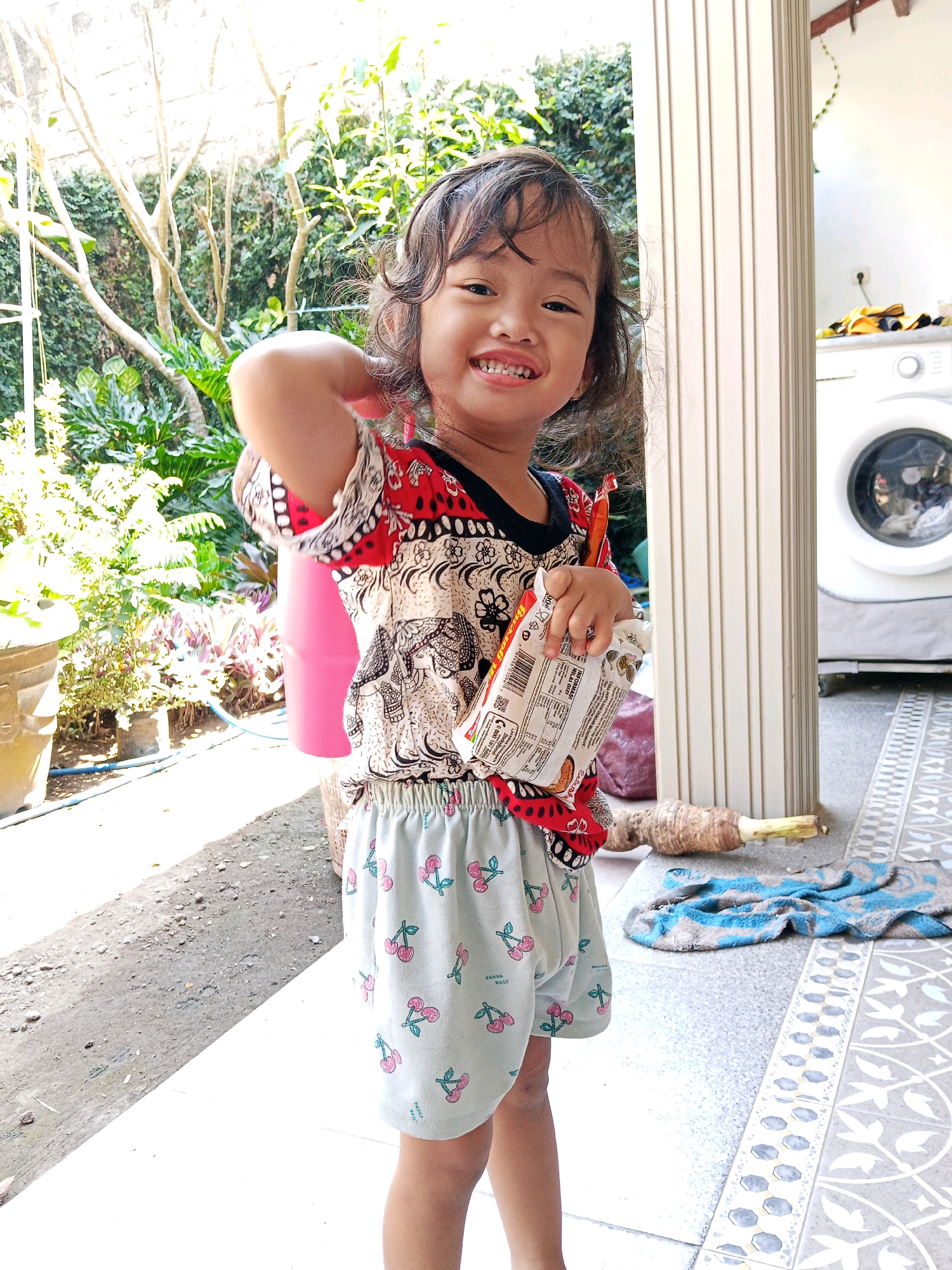 A young girl stands outdoors, smiling and holding a snack packet. She is wearing a patterned shirt with red, black, and white designs and light blue shorts with a cherry pattern. Behind her, lush greenery and trees are visible. To the right, a white washing machine stands near a pillar. The ground is paved with tiles and gravel. The scene appears to be a residential area in Gaprang, Indonesia.