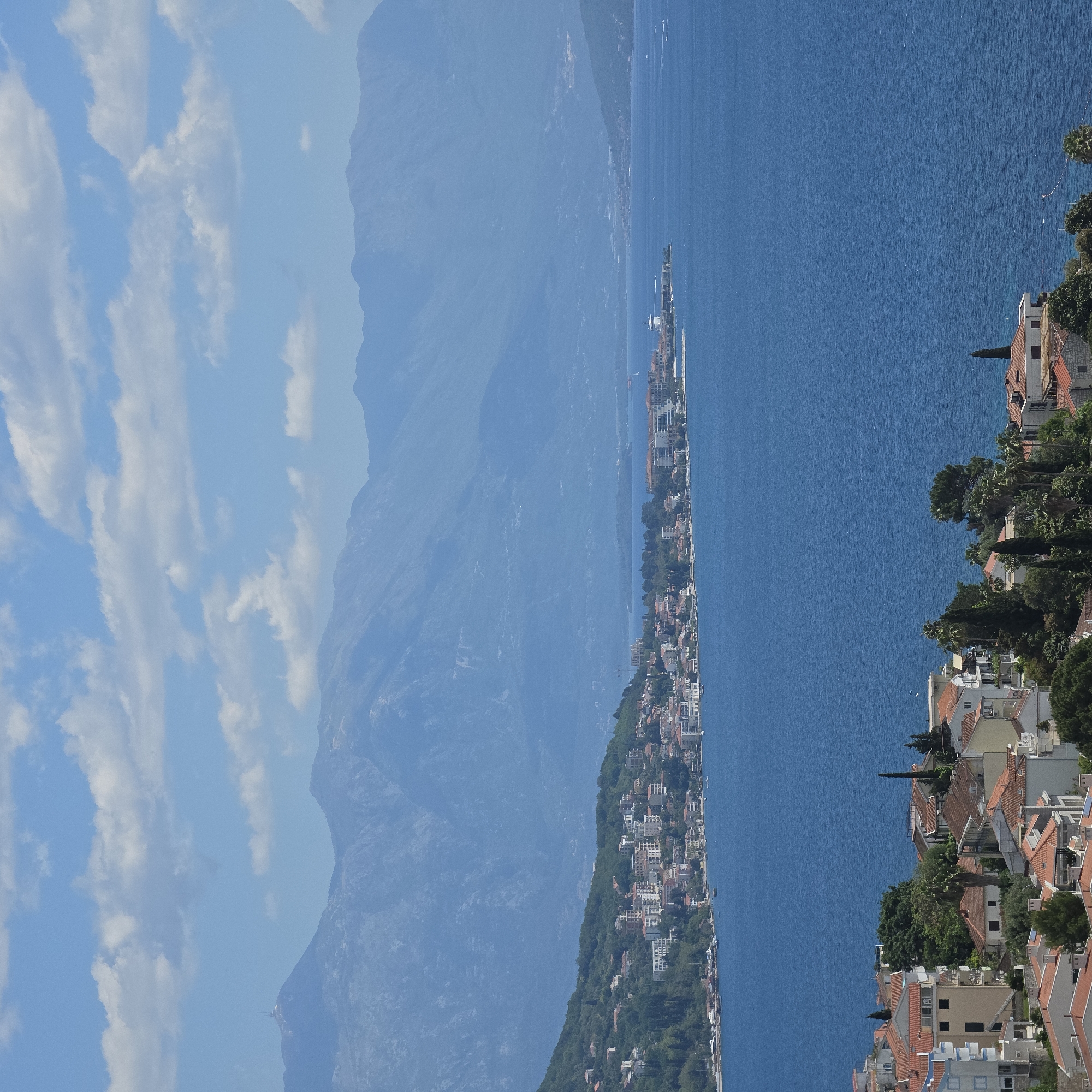 The image shows a panoramic view of a coastal landscape. The scene includes mountains shrouded in a hazy blue atmosphere and a body of water, likely a sea or bay. A narrow strip of land is populated with buildings, giving the impression of a settlement or coastal town. The architecture suggests a Mediterranean style with red-tiled roofs. Closer to the right side, there's a cluster of buildings amidst greenery, hinting at a mix of urban and natural elements. The sky is blue with scattered clouds. The overall impression is that of a scenic location, possibly a resort or historical area. This appears to be a view of Herceg Novi, Montenegro, with the long strip of land being part of the town stretching along the Bay of Kotor. The mountains in the background are typical of the Montenegrin landscape.