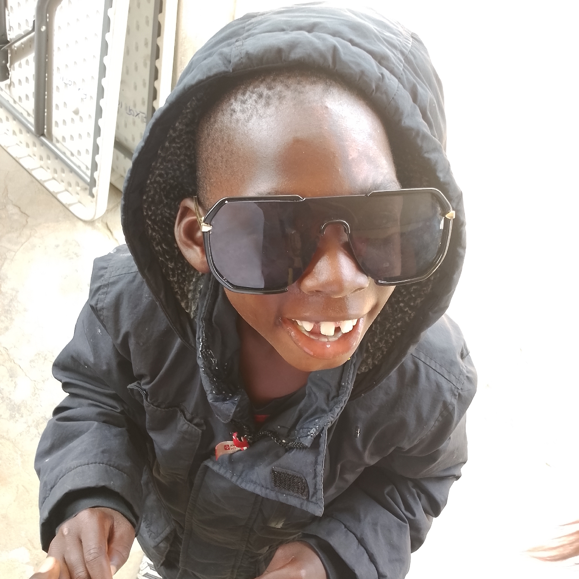 A young Black boy, wearing a black hooded jacket and large black sunglasses, smiles with his mouth open and teeth showing. He is outdoors, with a white surface to his left and a light-colored ground with visible cracks. The background is mostly white, suggesting a bright outdoor environment. The boy's hands are visible in the lower portion of the frame. There are no identifiable landmarks or contextual clues within the image to suggest a specific location in Ado, Nigeria.
