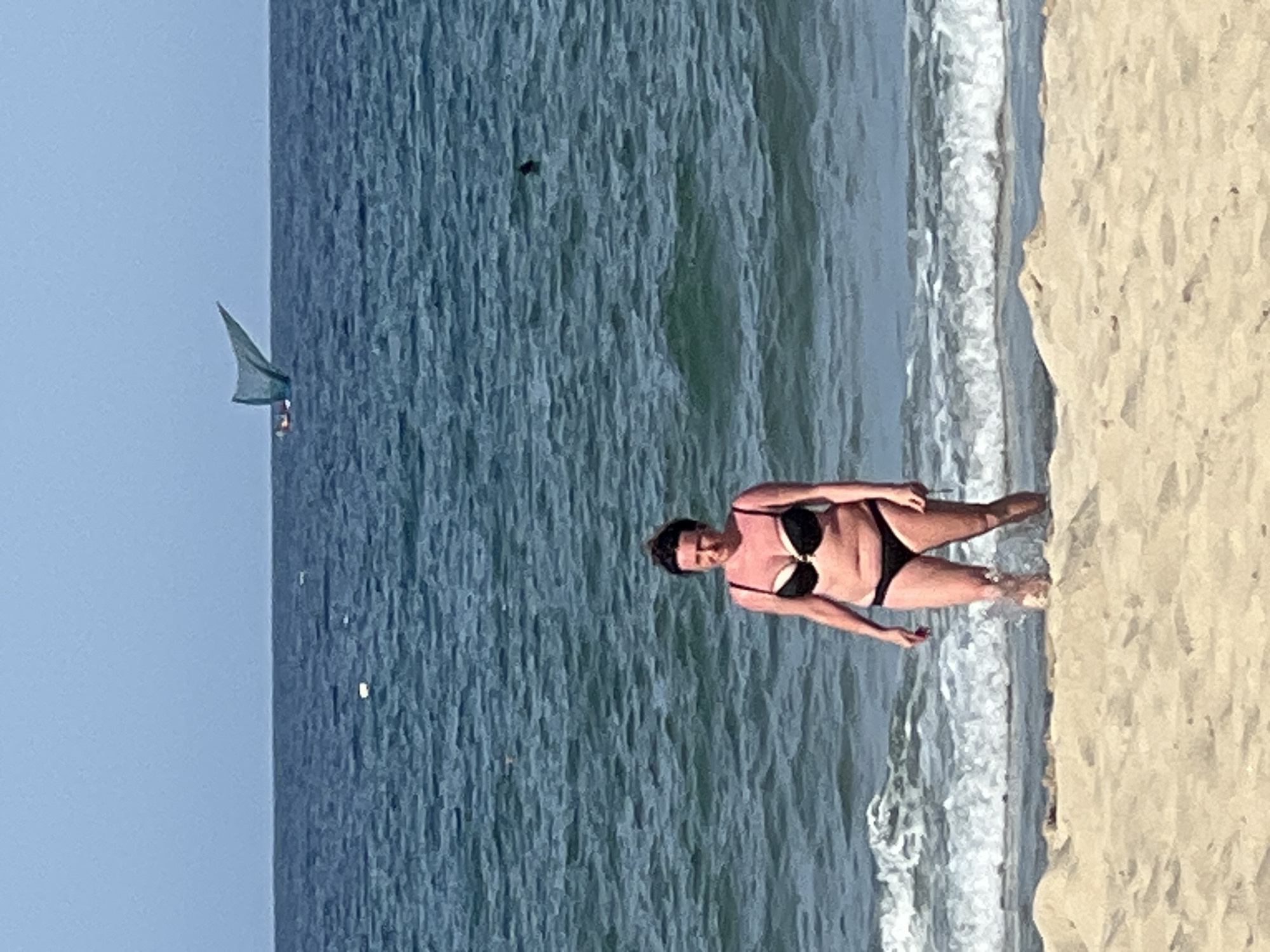A woman in a black bikini stands in the shallow water on a sandy beach. She has her hair pulled back and is looking at the camera. Behind her, the ocean stretches to the horizon, with a small boat with a triangular sail visible in the distance. The sky is clear and blue. This scene is likely located on one of the beaches of Da Nang, Vietnam, such as My Khe Beach or Non Nuoc Beach, which are known for their long stretches of sand and calm waters.