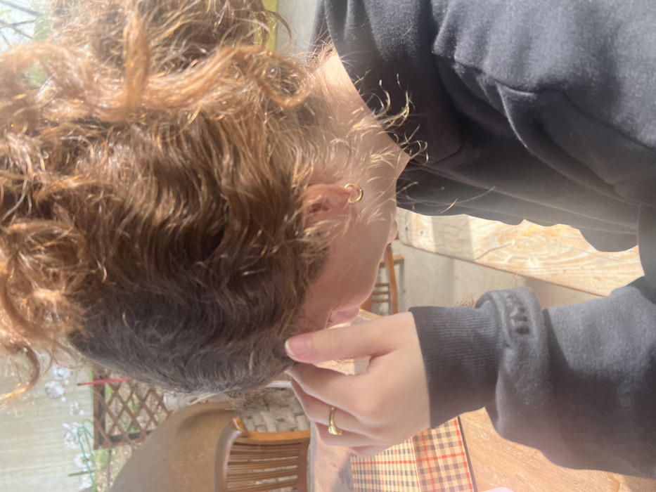 The image displays a close-up, side profile view of an individual, likely female, observed from the right, situated in an outdoor or semi-outdoor setting during daylight. The individual is facing towards the left of the frame.
The subject has wavy, light brown hair gathered in an updraft, possibly a messy bun or ponytail, with some loose strands framing the right ear and nape of the neck. They are wearing a dark-colored, long-sleeved hooded garment, consistent with a sweatshirt. A single, small, gold-colored hoop earring is visible in the right earlobe. A plain gold-colored band ring is worn on the ring finger of the right hand. The right hand is raised, with fingers gently pressed against the temple or side of the head, partially obscuring the right side of the face.
In the foreground, a light-colored wooden table or surface is present. A reddish-brown and beige checkered pattern, possibly a placemat or a printed design on the table, is visible to the left of the subject's hand.
The background features elements of an outdoor environment. A portion of a curved, dome-like structure, constructed of stone or brick with a lighter, possibly stucco or clay finish, is visible behind the subject. Adjacent to this structure, a wooden bench or chair with a slatted back is partially discernible. Further behind, a light-colored wooden trellis or fence is present, with hints of green foliage beyond it.
The scene is brightly illuminated by natural sunlight, casting highlights on the individual's hair and the wooden surfaces.
While the location is specified as Hof Ashkelon Regional Council, Israel, the visual elements within the image do not provide any specific identifying features or landmarks to forensically confirm this geographical detail. The scene depicts a generic outdoor leisure or dining area.