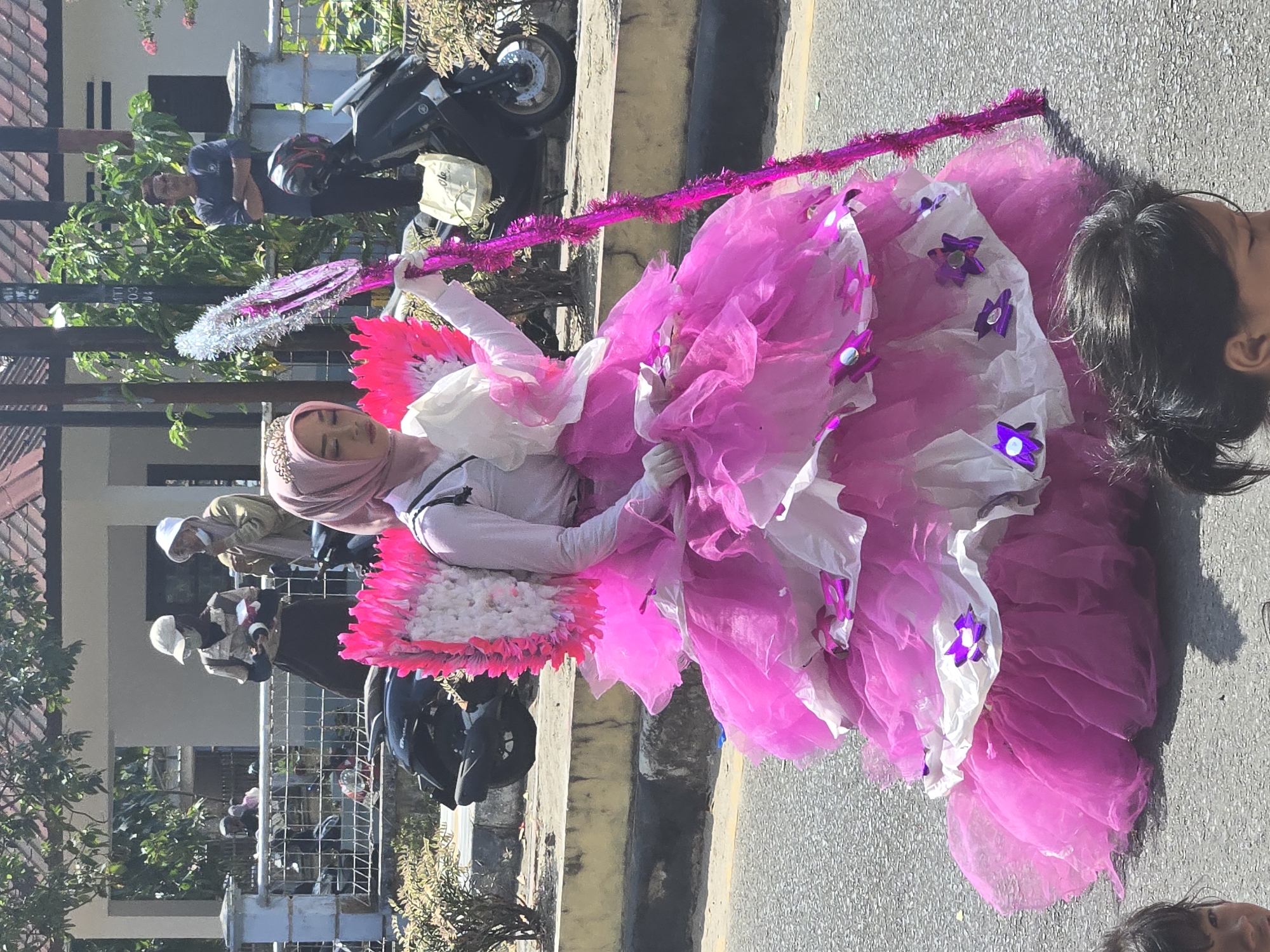In Sungailiat, Indonesia, a young woman in a pink and white tiered dress, adorned with pink feathered wings and a tiara, holds a sparkling pink staff while participating in a street parade. She wears a pink hijab and white gloves. Behind her, other people are visible, some watching the parade and others on motorcycles. The event appears to be taking place on a sunny day with palm trees and buildings in the background.