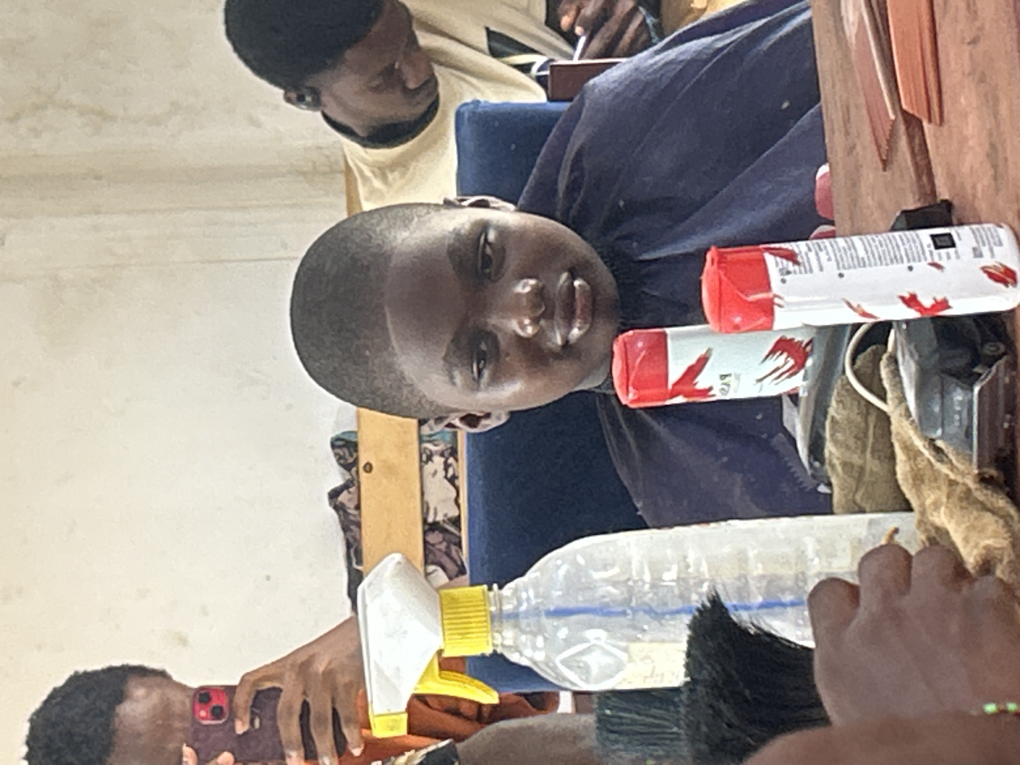 A young person with closely cropped hair is sitting in a blue barber chair, wearing a purple cape, likely at a barbershop. A person, presumably the barber, is visible in the background, standing. A water spray bottle and hair product are visible on a wooden surface next to the child. Another person can be seen taking a photo with their cell phone at the lower left edge. The specific city and location are not discernable from the image.