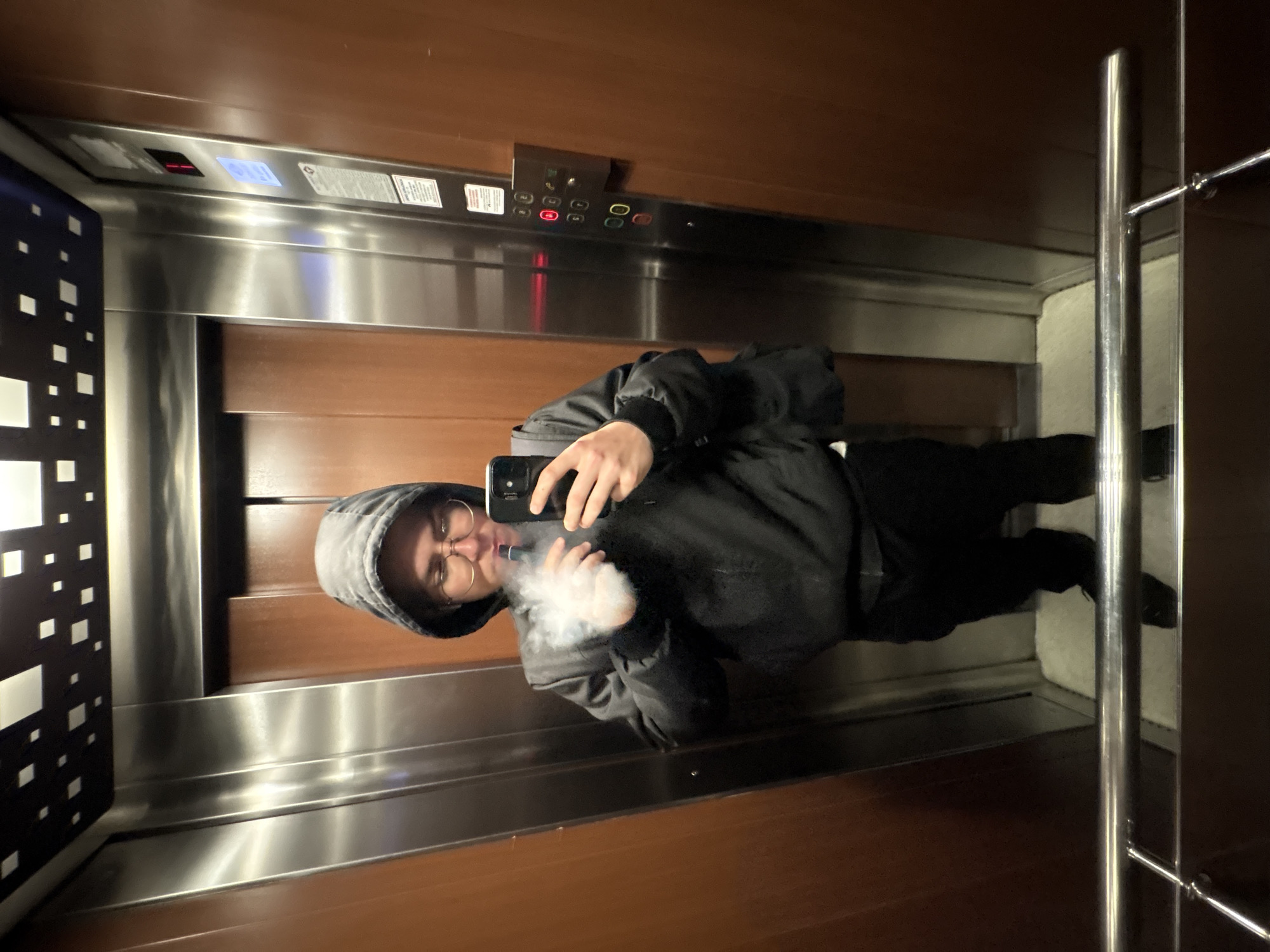 This image captures a person taking a selfie inside an elevator, which is located in Plovdiv, Bulgaria. The person, wearing a dark hooded jacket and round glasses, holds a black smartphone to take the picture while exhaling a large cloud of vapor from a vaping device held near their mouth.
The elevator's interior features a modern design, with walls composed of dark brown wooden panels and reflective brushed stainless steel. A metal handrail is visible on the right side. On the left, a decorative panel with a pattern of white squares on a dark background adds visual interest.
An elevator control panel is situated on the back wall, featuring several illuminated buttons, including red ones for floor selections (such as "00", "1", "2") and a yellow button. Above the control panel, a digital display shows "1" in red, indicating the current floor, along with a blue upward arrow. Several small informational or warning labels are affixed to the stainless steel panel; while most text is indistinct, one sign appears to contain a warning symbol and text, and another displays the number "1041". A light-colored mat is visible on the elevator floor. The lighting within the elevator is bright, creating reflections on the metallic surfaces. The person's facial expression is neutral, partially obscured by their glasses and the exhaled vapor.