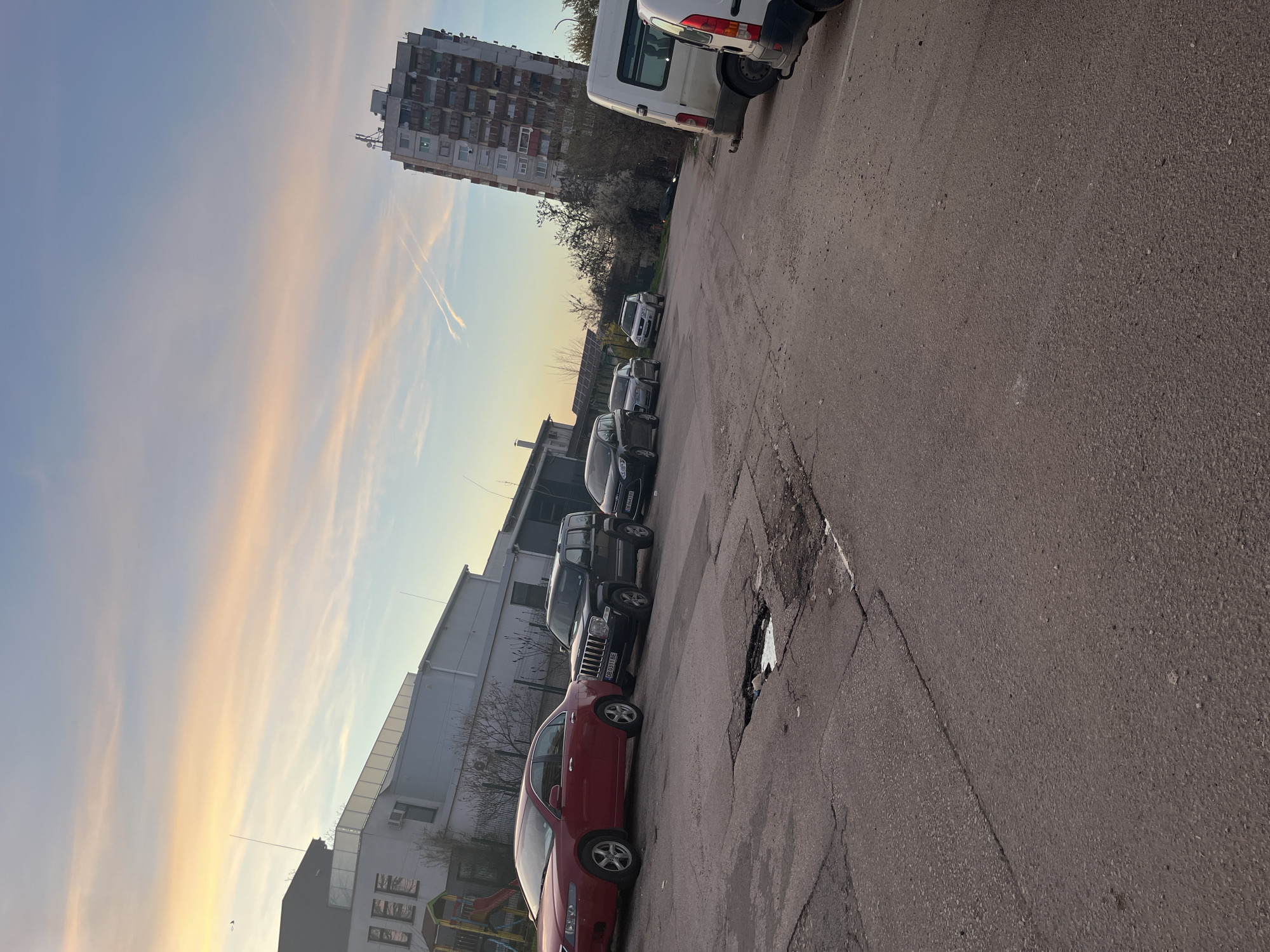 The image depicts a street scene in Lovech, Bulgaria. The perspective is angled, showing parked vehicles lining the side of the road. A red sedan is parked prominently at the lower edge of the image. Further down the street, a row of dark-colored cars and a white van are parked. A tall, multi-story apartment building is visible in the background. To the left, a lower-rise building is present. The sky is visible, with streaks of pink and blue suggesting either sunrise or sunset. The asphalt street shows signs of wear and tear.