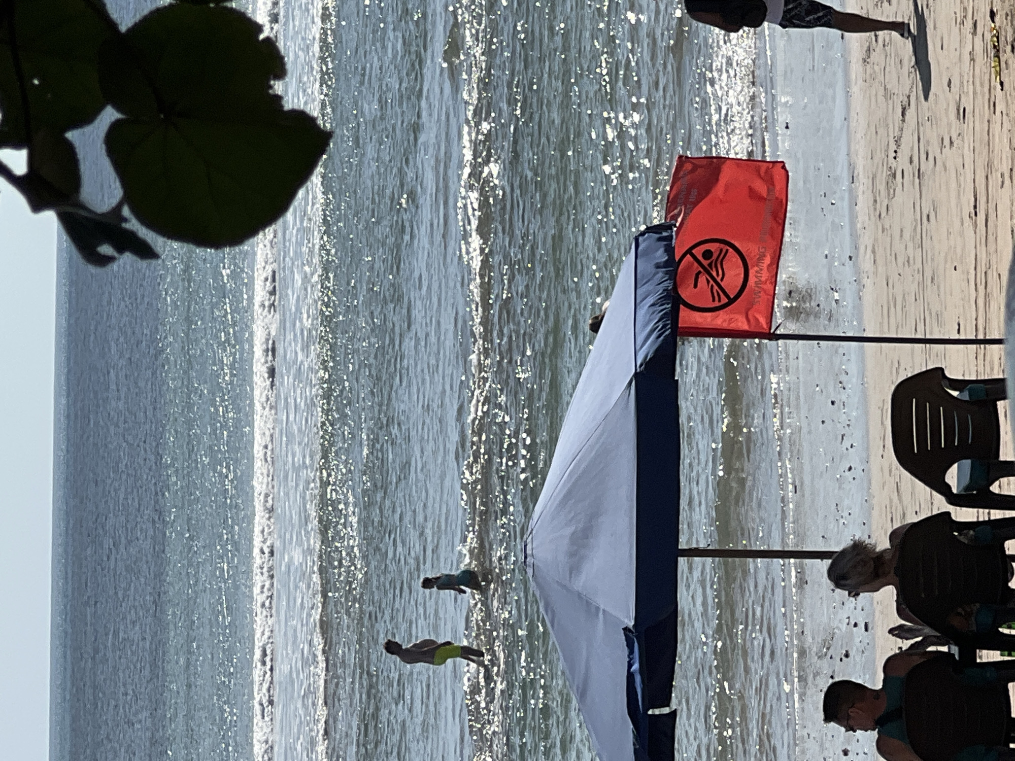 In Kuta, Indonesia, on a sunny day, two people are standing in the ocean with waves gently breaking around them. In the foreground, a large blue umbrella shades a section of the beach. A red flag with a prohibition symbol for swimming is planted nearby. Several people are seated in chairs on the sand, facing the ocean. The sky is clear and blue.