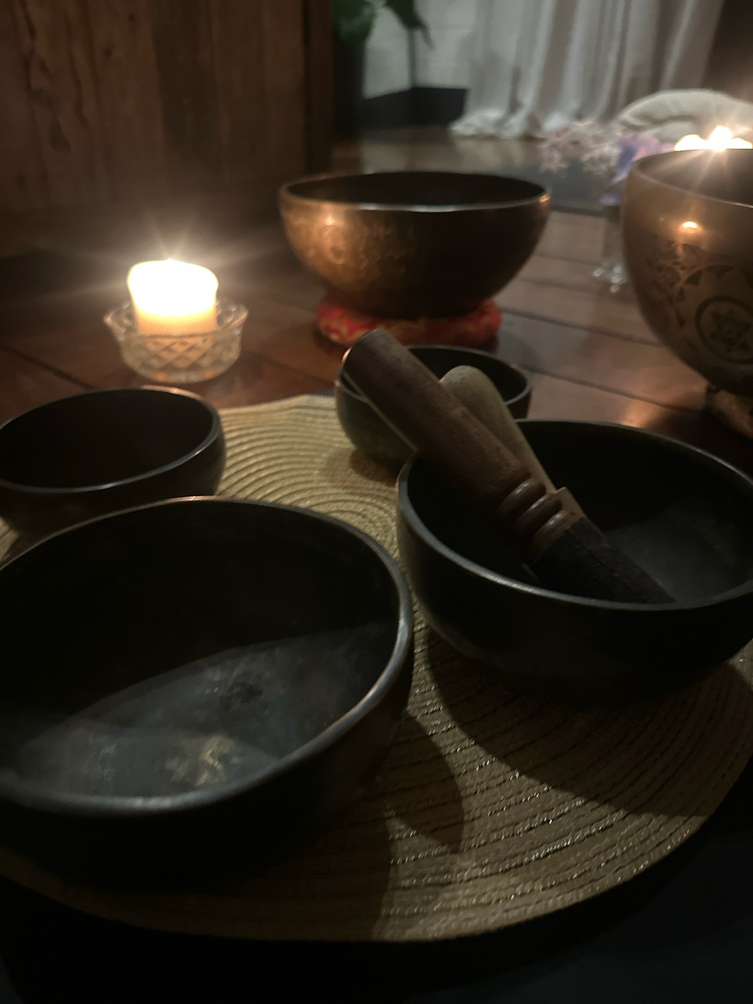 The image depicts a serene, dimly lit indoor setting, likely a room dedicated to relaxation or meditation. In the foreground, four dark, empty Tibetan singing bowls are arranged on a woven mat. One bowl contains a wooden mallet with a felted end. To the left and further back, a single lit white candle in a small glass holder casts a warm glow. In the background, larger bronze singing bowls are visible, one adorned with intricate carvings, resting on red cushions. The environment appears to be a floor or low table made of wood, with draped white curtains and hints of greenery in the distance, suggesting a peaceful, private space. The image does not contain any information indicating a location in Bellingen, Australia.