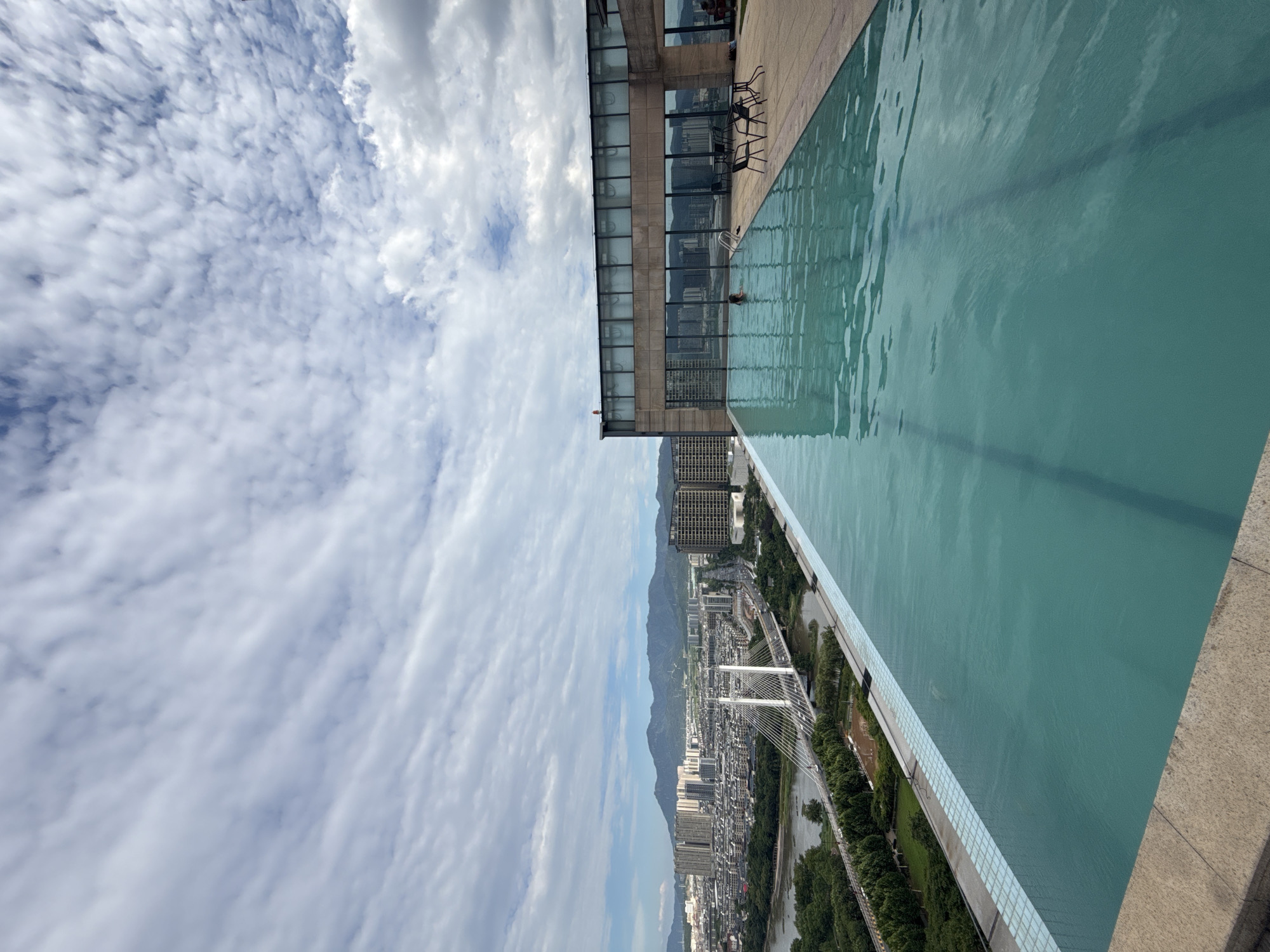 A high-angle view captures an infinity swimming pool on a rooftop, overlooking the city of Yiwu, China. The pool's turquoise water stretches towards the horizon, seamlessly blending with the sky. In the middle distance, a white cable-stayed bridge spans a river, connecting parts of the city. Buildings of various heights, including high-rises and residential blocks, form the urban landscape, with green trees lining the riverbanks. Distant mountains form a hazy backdrop against a sky filled with dramatic, layered clouds. A person is visible in the pool, and a few tables and chairs are set up on the pool deck. The scene conveys a sense of urban tranquility and expansive city views.