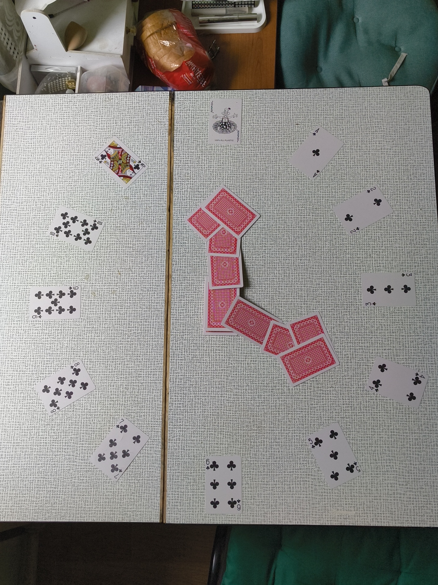 The image shows a close-up, high-angle view of a table set up for a card game, likely in a domestic setting. The table surface has a textured, light-colored pattern. A variety of playing cards are spread across the table, with some face up and some face down.
On the left side of the table, several face-up club cards are visible, including a 10, an 8, and a 6. A Queen of Hearts is also displayed prominently on the left. On the right side, there are more face-up club cards, including a 4, a 2, a 3, and another 4. In the center of the table, a cascade of red-backed cards is arranged in a diagonal line, suggesting they might be part of a draw pile or a tableau.
A single joker card, marked "100% ALL PLASTIC," is positioned near the top center of the table. In the background, partially visible, are a loaf of bread in plastic packaging, a white storage container, and a portion of a teal-colored chair. The lighting suggests an indoor setting, likely during the daytime. There are no people visible in the image. The overall impression is a paused moment in a card game.