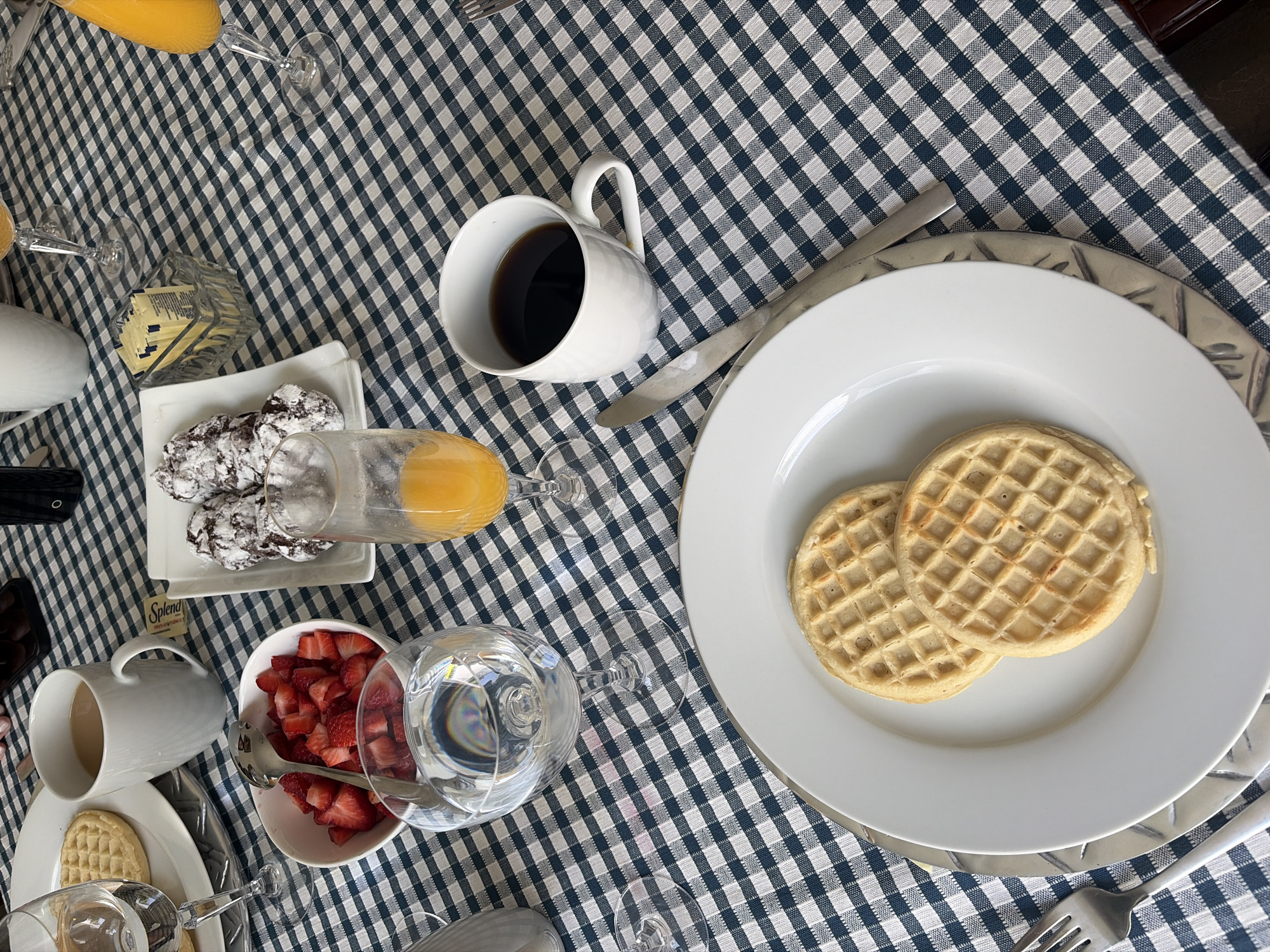 A tabletop scene is presented on a blue and white checkered tablecloth. At the top right is a white plate holding two waffles, accompanied by silverware to its right. Directly to the left, a white mug holds a dark liquid, possibly coffee. Further left, a small white dish displays several small, powdered chocolate cookies, with a glass of orange juice adjacent. Towards the bottom left, another white mug contains a lighter-colored beverage, and a white bowl is filled with sliced strawberries, a spoon resting in the bowl. A tall glass of water stands nearby. The location is unknown.