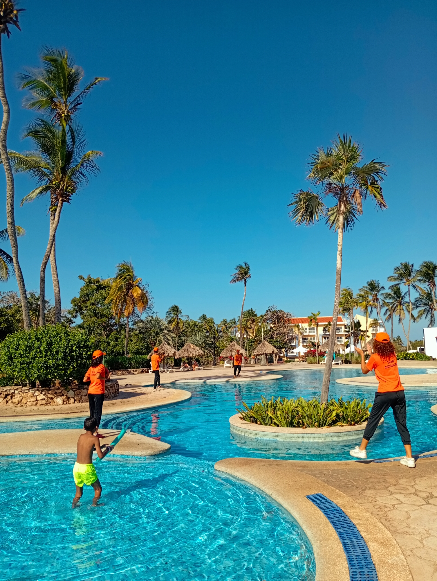 At a resort pool in Playa El Agua, Venezuela, a young boy wearing neon green swim trunks stands in the clear, turquoise water, holding a long, blue pool noodle. Around the pool, on tan-colored walkways, are several adults wearing matching bright orange t-shirts and hats. The shirts have black writing, partially obscured. One adult stands near the boy, while others are positioned further away around the pool. Palm trees are scattered throughout the scene, framing the bright blue sky. Thatched-roof structures can be seen in the background, along with a multi-story building.