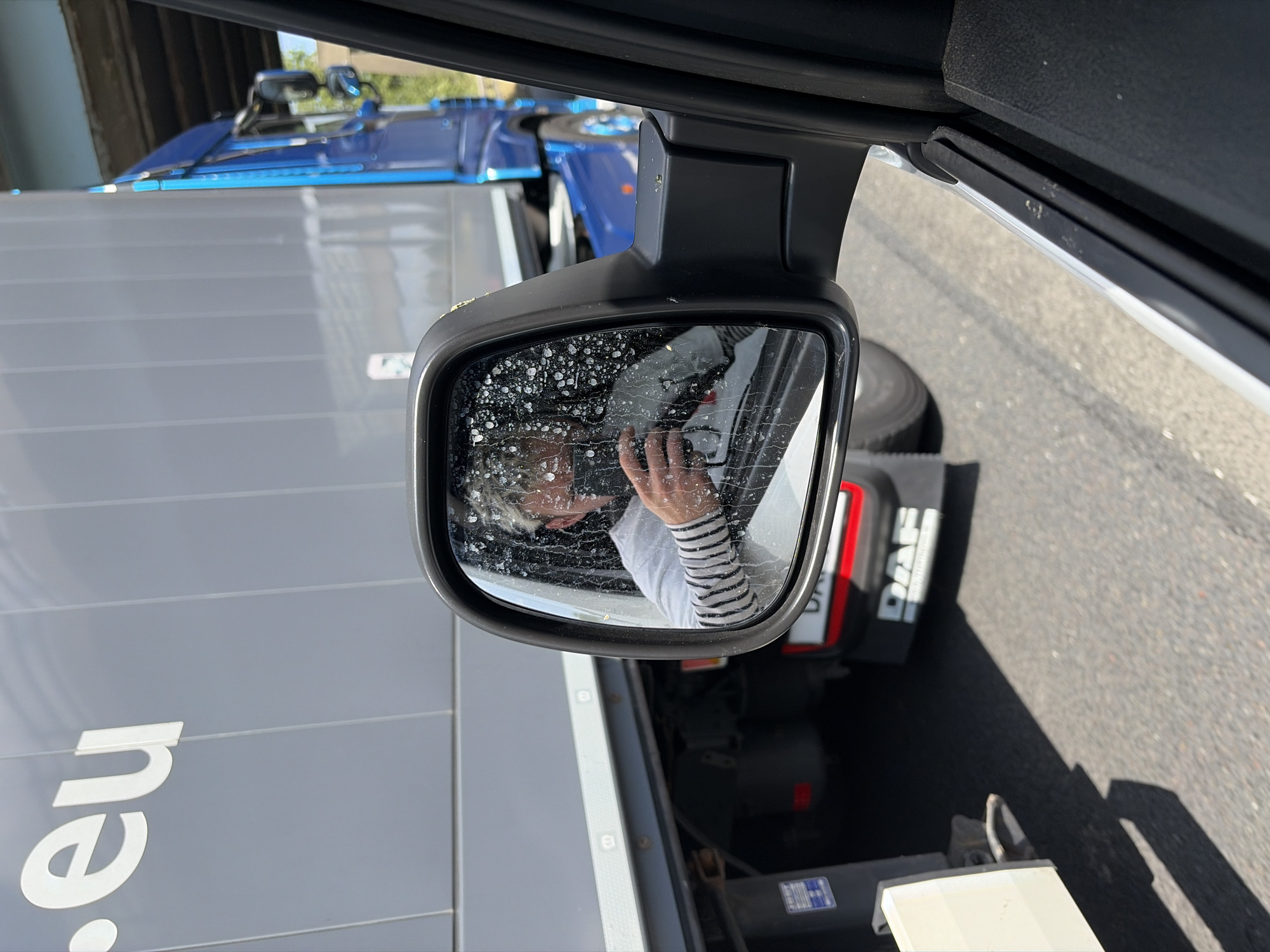 The image shows a person taking a selfie in a side-view mirror of a truck. The person, with short blonde hair and wearing a striped shirt, is holding up a phone to capture the reflection. The mirror is dirty with water spots and streaks, obscuring some of the reflection.
The setting appears to be a roadside or industrial area, with a large grey truck trailer in the foreground and a blue truck in the background. The trailer has white text that is partially visible, reading ".eu". The blue truck has "DAF" branding visible on its side. The ground is asphalt, suggesting a road or parking lot. Sunlight indicates it is daytime, likely midday given the brightness. The overall mood is casual, as indicated by the selfie. No specific emotions are discernible beyond the act of taking a photograph. The location context of Rochdale, United Kingdom is noted, but not directly evident from the visual elements within the image itself.