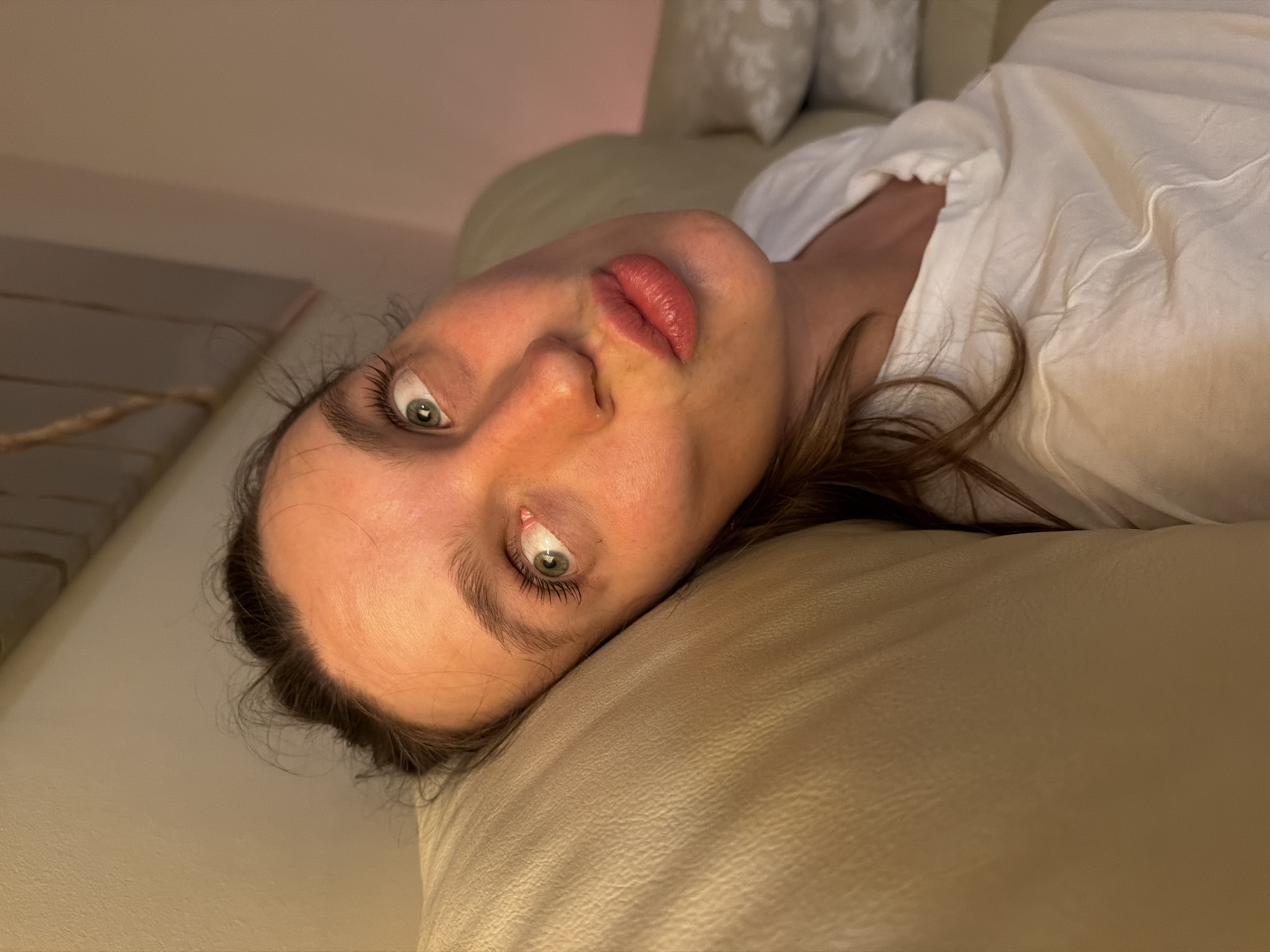 A close-up, rotated portrait shows a person, likely a young adult female, lying down indoors. The individual has fair skin, prominent green eyes, and light brown hair, which is casually spread around her head. She is looking directly into the camera with a direct and curious gaze. Her lips are slightly parted, revealing a soft red or pink lipstick.
She is resting her head on a light-colored, possibly beige or cream-toned, upholstered surface, which appears to be a couch or armchair. She is wearing a light-colored, likely white, loose-fitting top or shirt. The background is softly blurred, showing other light-toned furniture elements, possibly pillows.
The lighting is soft and warm, suggesting an indoor setting during the evening or night. The image is rotated approximately 90 degrees counter-clockwise, with the person's head towards the upper left of the frame. The overall atmosphere is intimate and relaxed. There is no visible text in the image. The context given for the location is Budva, Montenegro.
