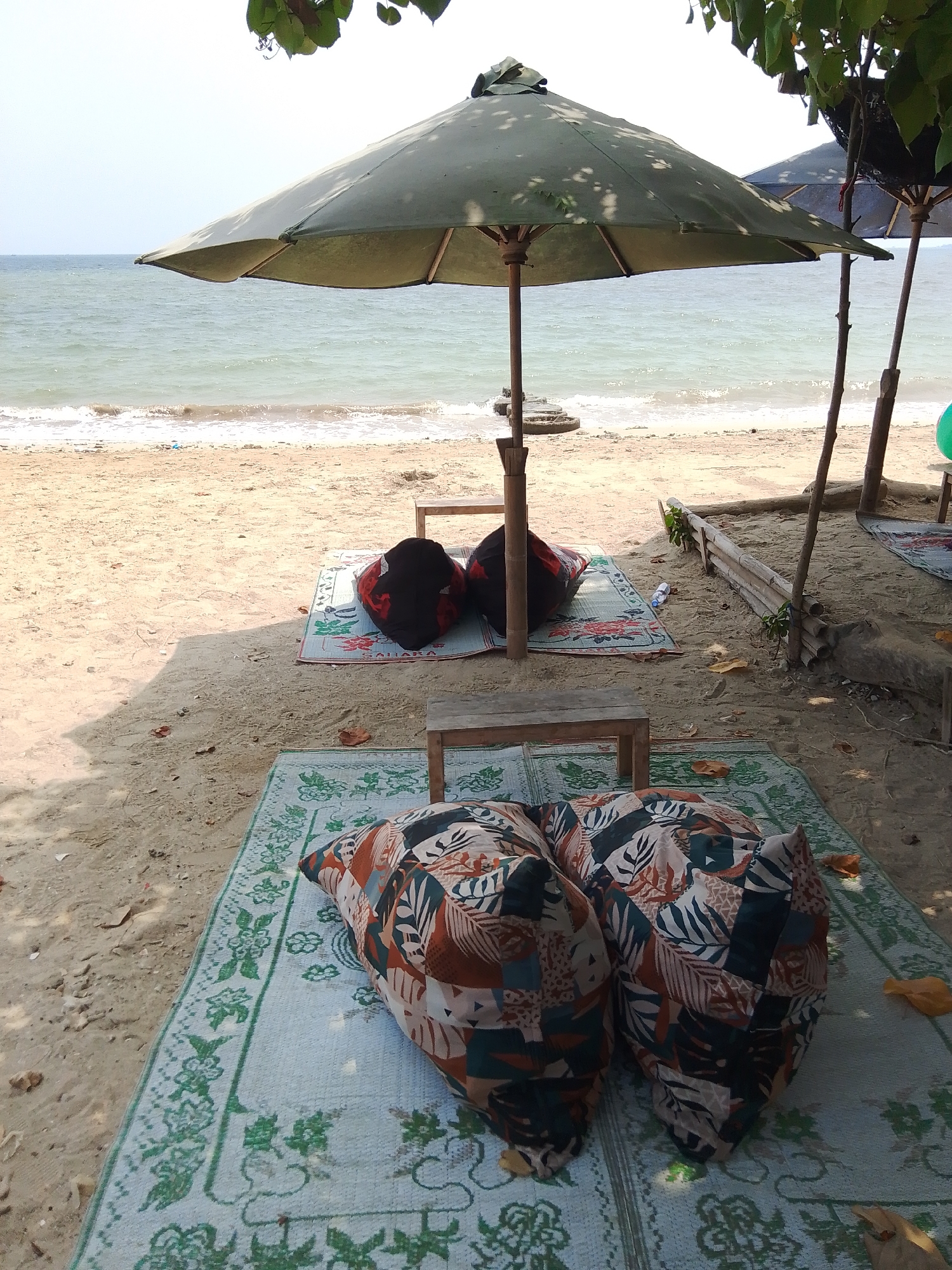 The image depicts a beach scene in Tegalsambi, Indonesia, featuring a relaxed seating area under a large green umbrella. Two patterned floor cushions are visible in the foreground, placed on a light blue mat with a green floral pattern. Behind these, another mat holds two dark-colored cushions. The beach itself is sandy, with the gentle waves of the ocean visible in the background. Trees with green leaves provide shade, casting shadows on the sand. The overall impression is a peaceful, outdoor relaxation spot.