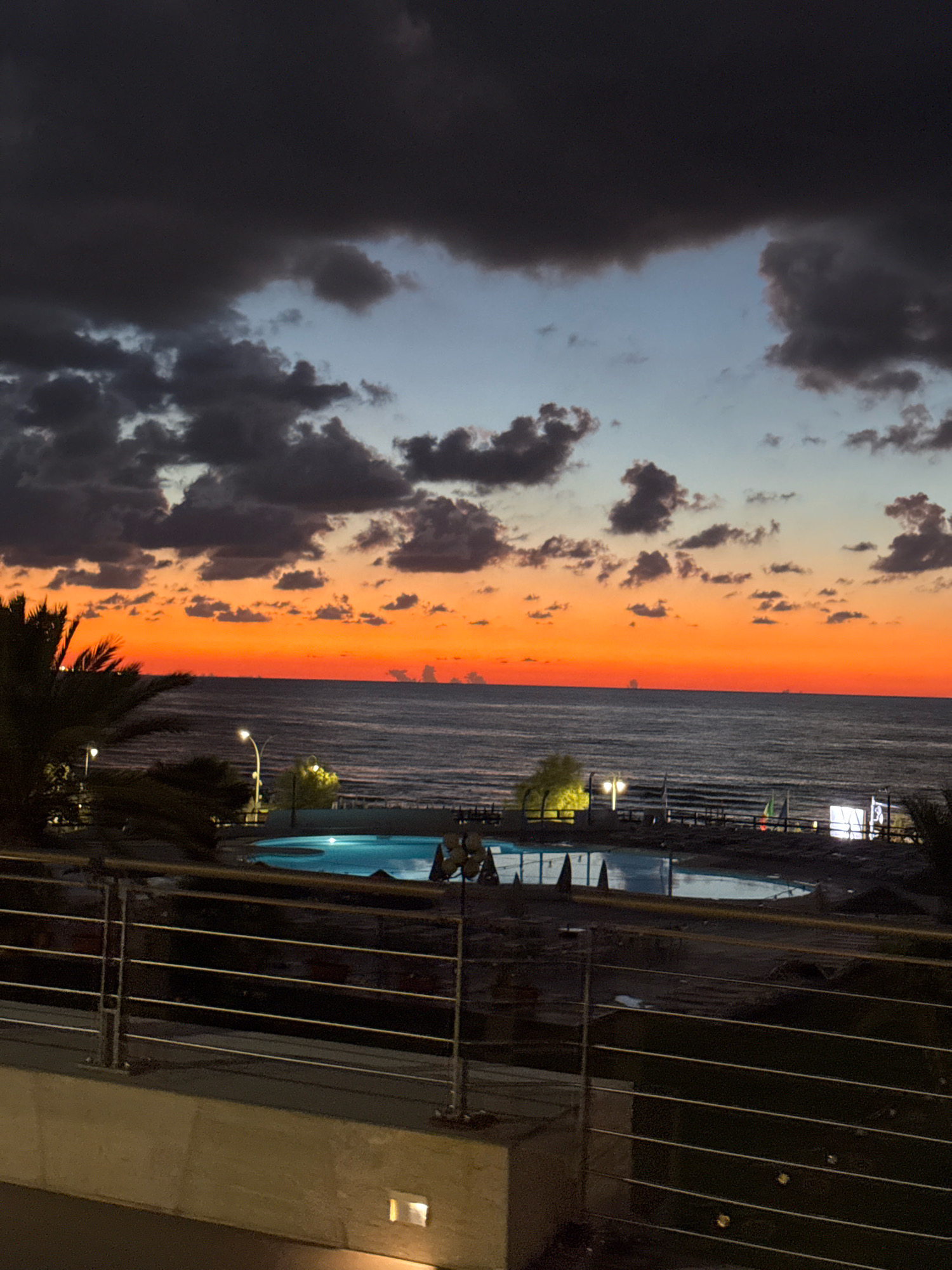The image depicts a sunset over the sea in Isola delle Femmine, Italy. The sky is a vibrant gradient of orange, transitioning to blue and then to dark clouds at the top. The sea is calm, with a visible horizon line. In the foreground, a swimming pool with illuminated blue water is visible, surrounded by dark ground. A metal railing with horizontal bars runs across the bottom of the image, separating the viewer from the scene. Palm fronds are visible on the left side of the frame. The overall atmosphere is serene and twilight.