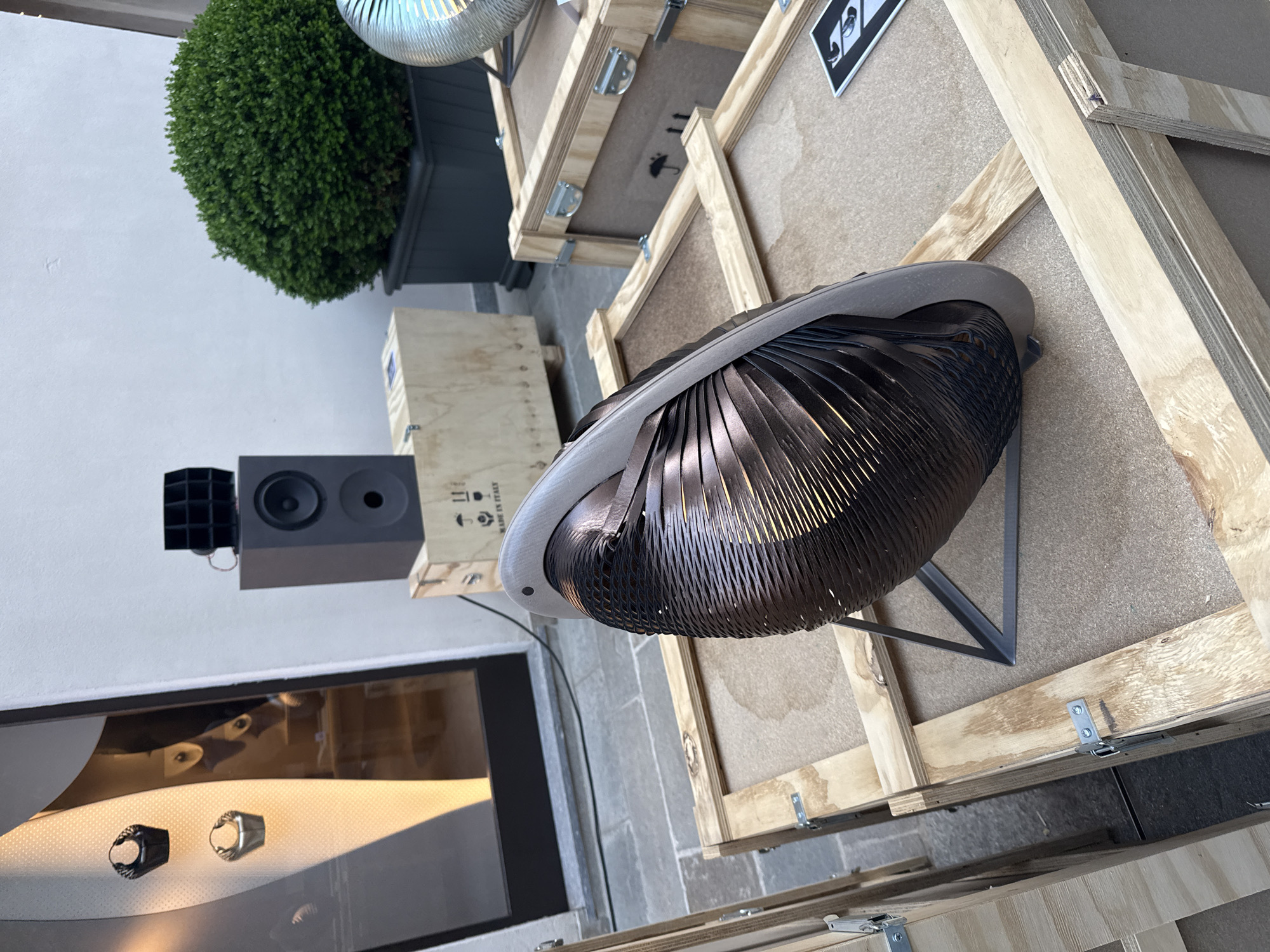 This media file captures an outdoor or semi-outdoor exhibition space, likely in Milan, featuring a curated display of objects under natural daylight. The central focus is a large, sculptural or decorative object, possibly a speaker or light fixture, composed of numerous dark, metallic, ribbon-like strips forming a spherical, open-weave structure. It has a lighter grey or beige rim and rests on a minimalist dark metal stand.
Around this central piece, several wooden shipping crates are visible. Some appear closed, while others are partially open or stacked. On one crate, the text "MADE IN ITALY" is partially legible, along with international shipping symbols such as "THIS WAY UP" arrows, a fragile glass symbol, and an umbrella indicating "keep dry." A small black and white print, possibly an artwork, featuring two stylized figures looking upwards, is affixed to one crate.
In the background, a dark grey, rectangular speaker box with a circular driver and a port is visible, accompanied by a smaller, black, multi-chambered horn-like speaker component. To the upper left, a vibrant green, spherical topiary plant sits in a dark planter against a plain white wall. The ground is paved with grey and tan stones. In the lower left, a large, dark, reflective surface, possibly a window or display case, shows reflections of the environment and two small, ornate ring-like objects, one dark and one metallic silver. The overall scene suggests an artistic or product display, possibly at an outdoor event or gallery space. No people are present, and the time of day appears to be daytime.