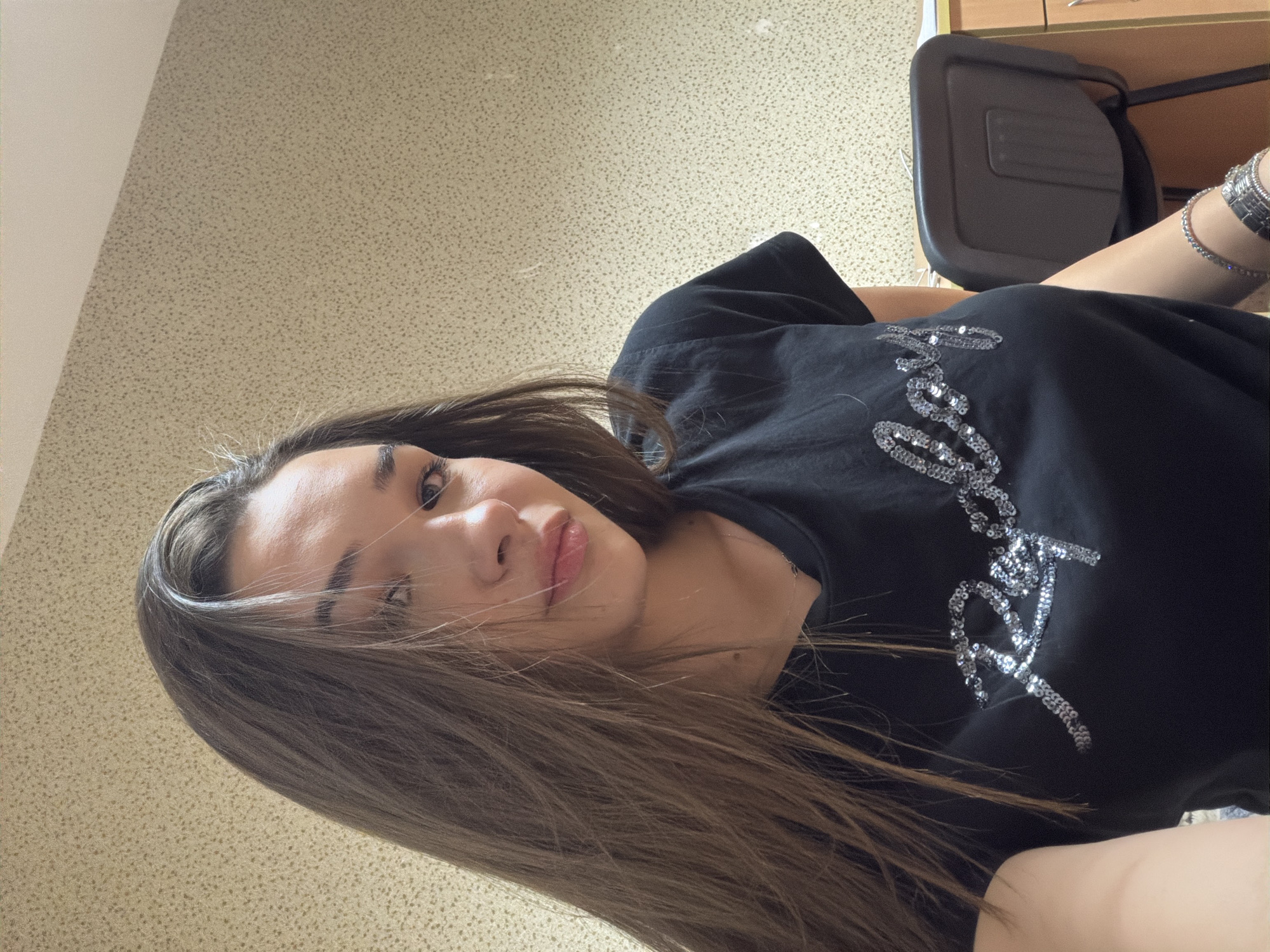 A young woman with long, brown hair is pictured from a low angle. She is wearing a black t-shirt with a silver, sequined design on the front. She has bracelets on her right wrist. She appears to be indoors, sitting in front of a beige-toned wall with a speckled pattern. A black chair is visible in the upper right of the frame, partially obscured by a wooden cabinet. There is no indication of the location being in the city of Sofia, Bulgaria.