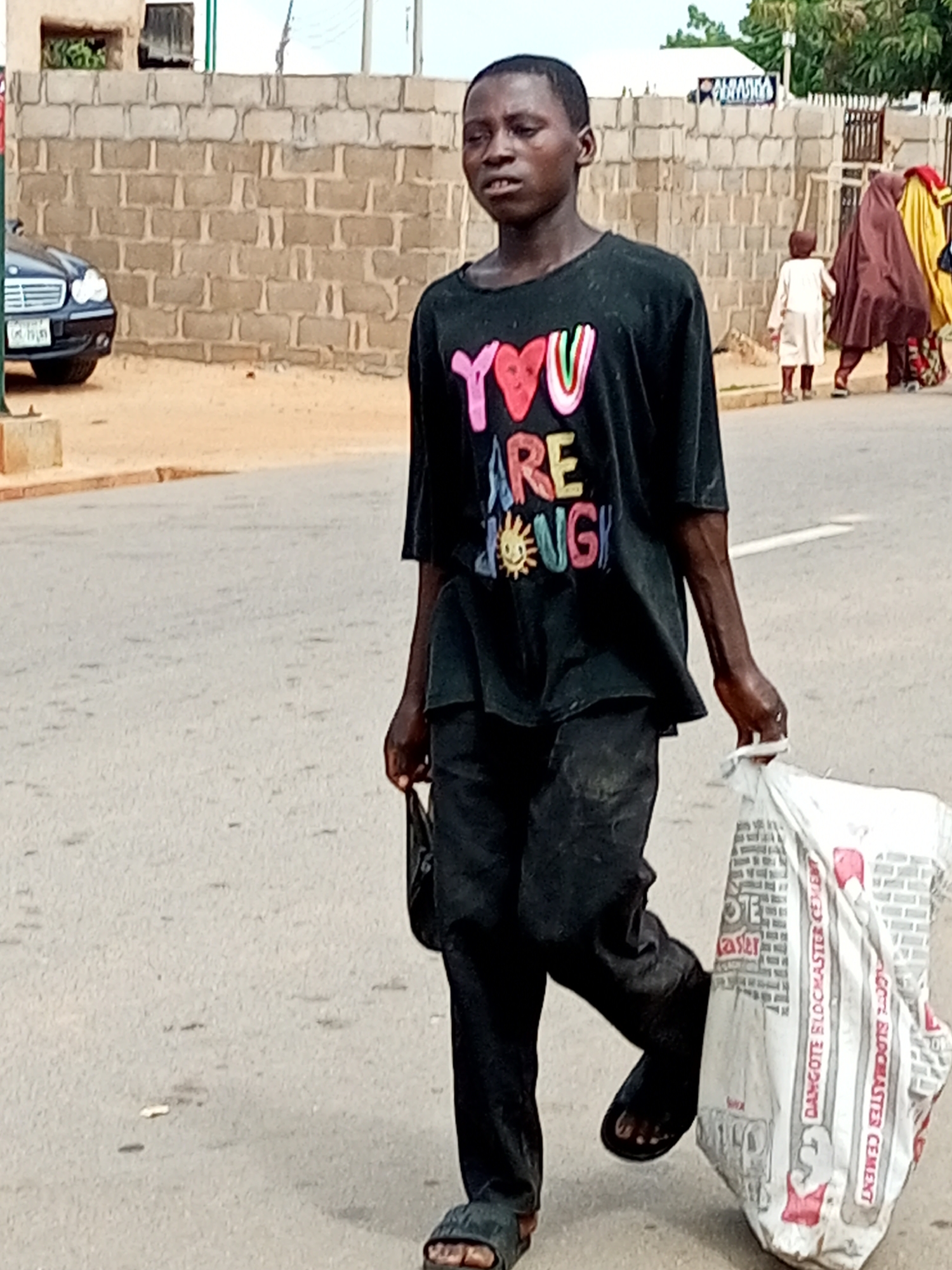 A young Black male walks across a paved road, carrying a large white bag of cement in his right hand and a small black bag in his left. He wears a black t-shirt with a colorful graphic on the front that reads "YOU ARE ENOUGH" with a sun wearing sunglasses. His pants are also black, and he wears dark-colored sandals. The person appears to be walking from left to right in the frame.
In the background, a brick wall lines the street, with a dark-colored Mercedes-Benz sedan parked to the left. Further down the road, two women in traditional Islamic attire, one in a brown hijab and the other in a yellow hijab, are walking away from the camera. The scene appears to be set in a town or city, with buildings and fencing visible in the distance. The lighting suggests it could be daytime, possibly late afternoon given the shadows. The overall atmosphere is that of everyday life in a Nigerian town, with people going about their business. The prominent text on the t-shirt suggests a message of self-acceptance.