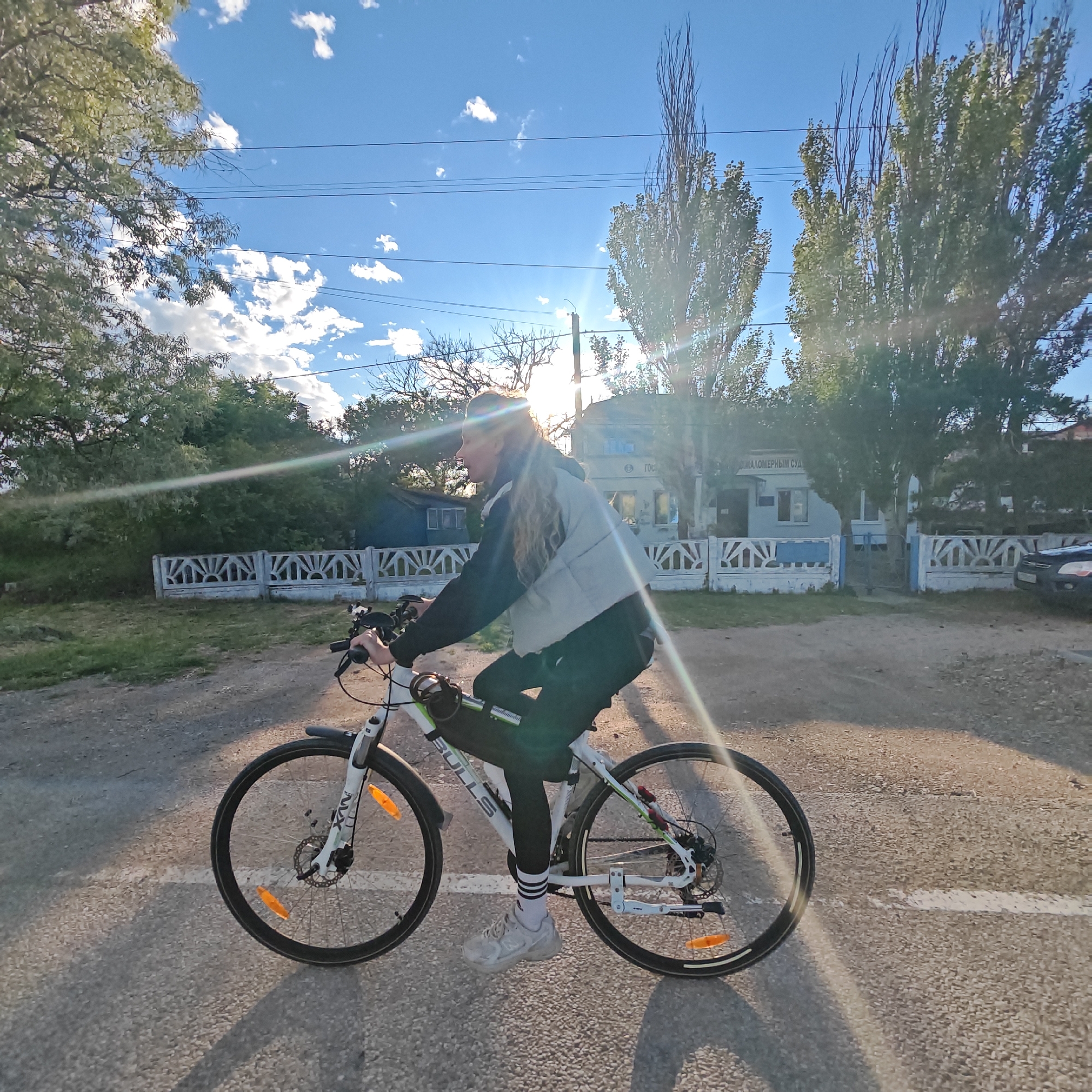 An outdoor, sun-drenched scene captures a person riding a white "BULLS" brand bicycle on a paved or dirt road in Мысовское сельское поселение, Ukraine. The individual, with long, light hair, is wearing a light grey puffer vest over a dark long-sleeved top, dark pants, and white-striped socks with light sneakers. They are looking ahead, towards the left of the frame, engaged in a casual bike ride.
The setting appears to be a rural or suburban area. A white line is visible on the ground beneath the bike. The strong sun, positioned behind the rider's head, creates significant lens flare and long shadows, indicating either early morning or late afternoon. The sky is bright blue with scattered white clouds, and power lines crisscross overhead.
In the background, lush green trees and some bare-branched trees line the path. Behind a decorative white and blue fence, a light-colored building with dark windows is visible. The building's facade features Cyrillic text clearly reading "МЫСОВСКОЕ СЕЛЬСКИЙ СУД" (Mysovskoe Rural Court). A dark-colored car is partially visible parked near the building on the right. The bicycle is equipped with "MXN" branded front suspension, disc brakes, and orange reflectors on the wheels.