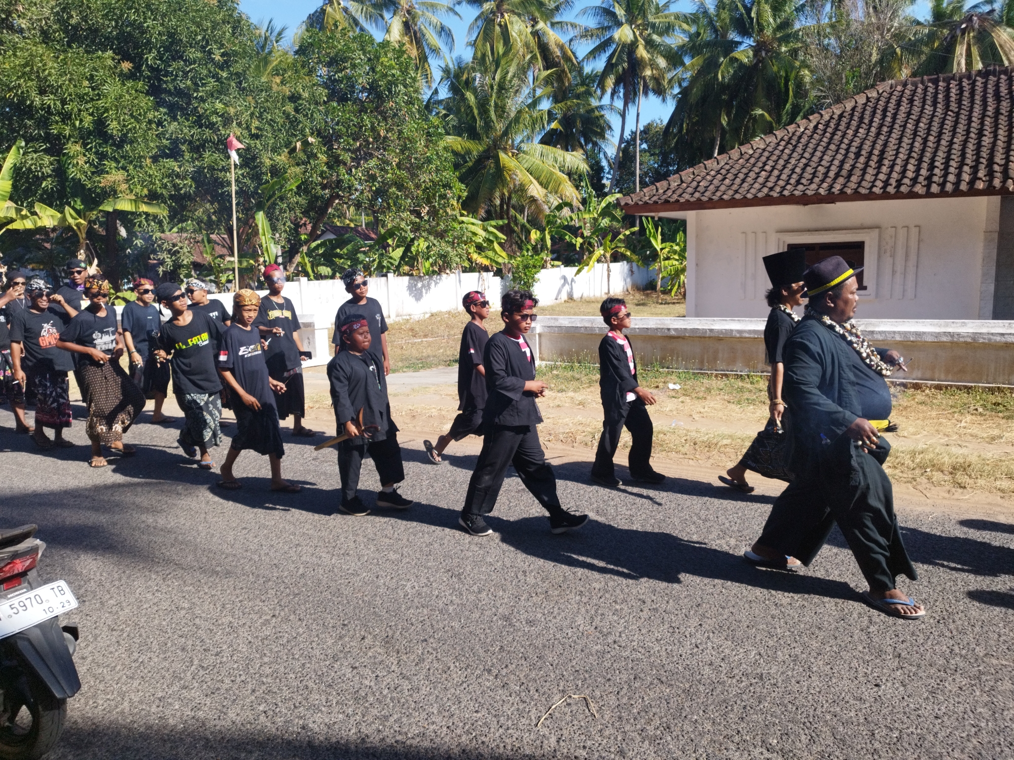 The image shows a group of people walking down a paved road in what appears to be a procession. The group is diverse in age, with many young individuals, likely children and teenagers, and some adults. They are dressed in dark clothing, with many wearing black shirts and pants. Some individuals are wearing headbands, and a few are wearing traditional hats. The environment is tropical, with lush green trees and palm trees in the background. A white building with a tiled roof is visible on the right side of the image, and a small Indonesian flag is planted on a pole near the trees. The road surface is asphalt, and there are shadows cast by the people and trees. A motorcycle is partially visible on the left side of the road, with a license plate reading "5970 TB 10.29". The scene suggests a cultural event or ceremony taking place in Andulang, Indonesia.