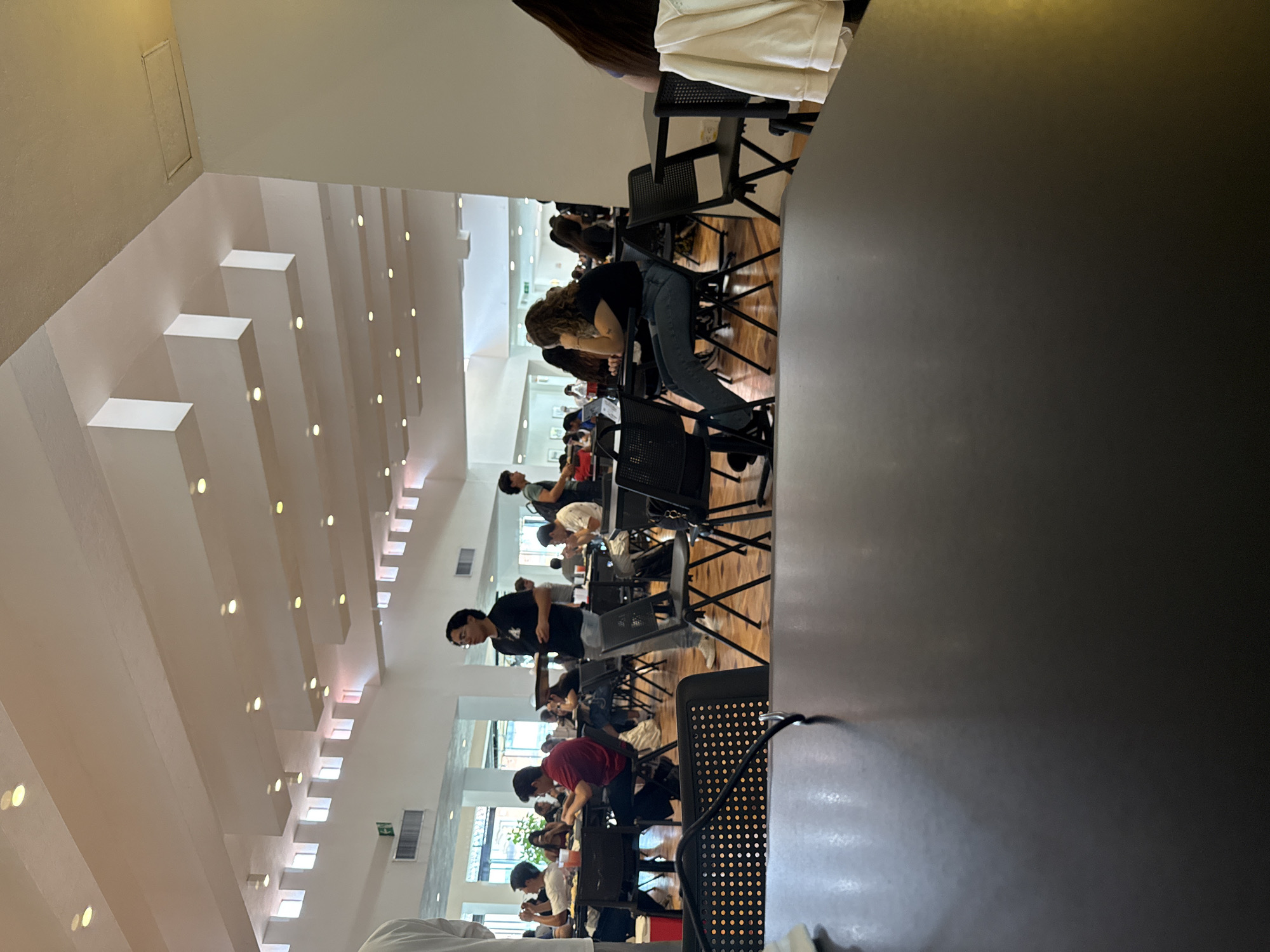 A well-lit interior space in San Andrés Cholula, Mexico, is populated by numerous individuals seated at tables. The architecture features a high ceiling with a repeating pattern of rectangular light fixtures. Many of the seated people appear to be studying or working at their desks, with black chairs at each table. The floor is wooden. Some individuals are wearing black shirts and some are wearing red shirts.