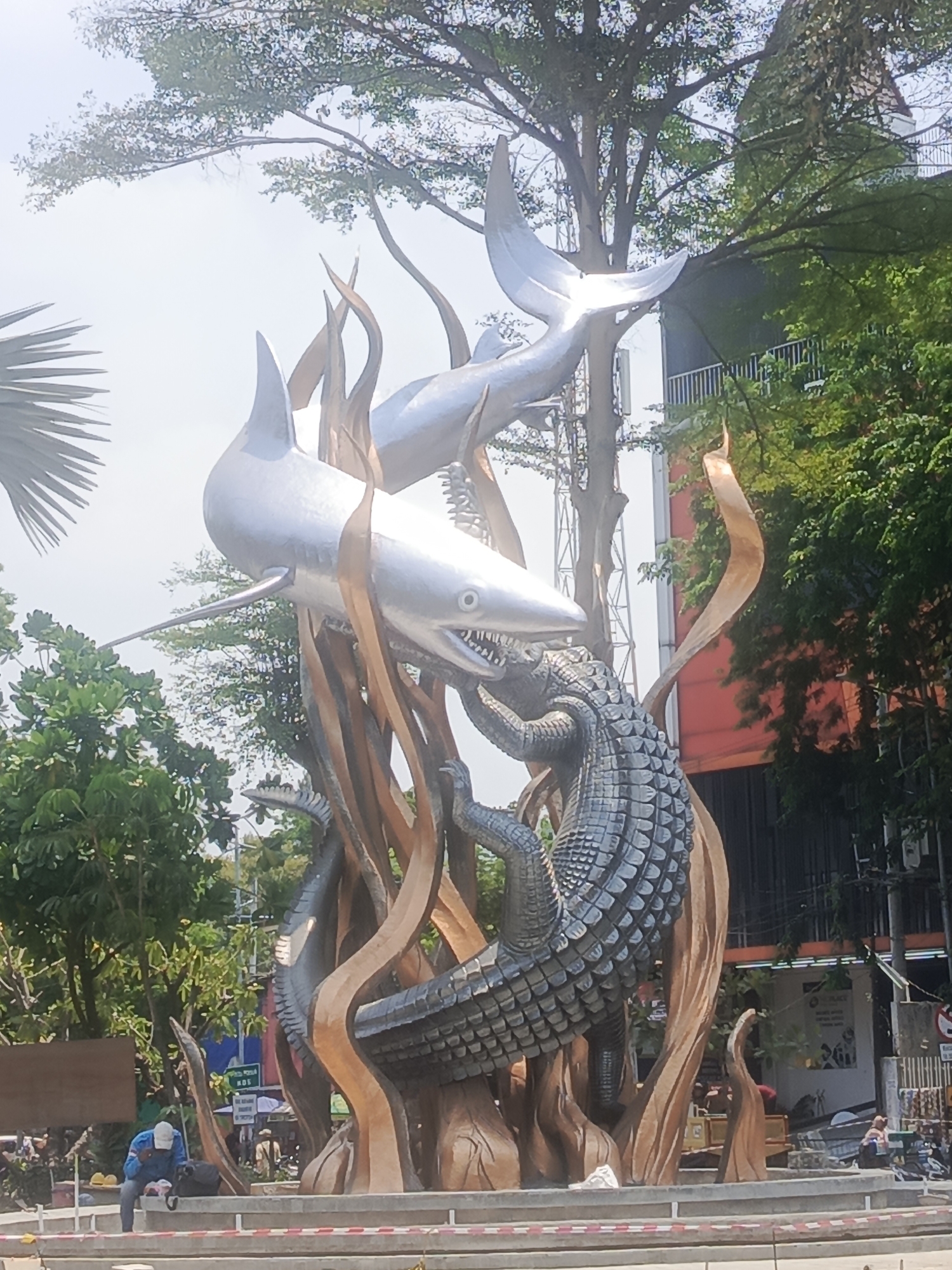 The image captures the iconic "Sura dan Baya" (Shark and Crocodile) statue in Surabaya, Indonesia, a significant landmark representing the city's legend. The sculpture depicts a fierce battle between a shark and a crocodile, set against a backdrop of lush green trees and a bright sky. The metallic sheen of the shark contrasts with the textured, dark scales of the crocodile. In the foreground, a person wearing a blue shirt and a white cap is sitting on a ledge, facing away from the camera. Various urban elements, including buildings and street signs, are visible in the background, indicating an urban environment. The scene appears to be a public space, likely a park or square, given the presence of the monumental sculpture and surrounding greenery.