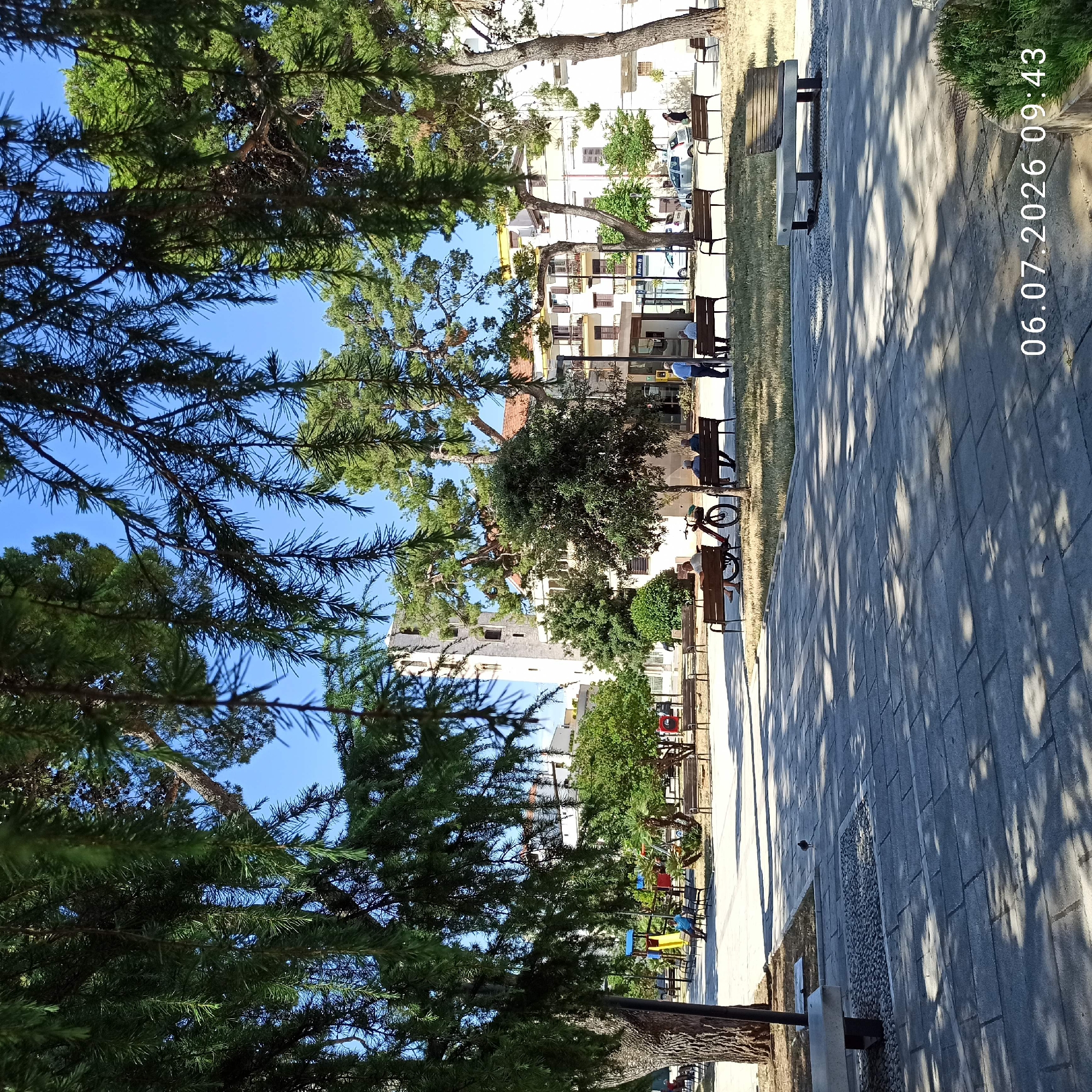 The image captures a sunlit public square in Ruvo di Puglia, Italy, on July 6, 2026, at 09:43 AM. The foreground features a paved walkway with a dappled shadow pattern cast by overhead trees. A single small bird stands on the pavement in the mid-ground. To the left, a bench is visible, and further back, a children's playground with colorful slides is present. Several individuals are seated on park benches throughout the square, some interacting and others observing their surroundings. A bicycle is parked near one of the benches. In the background, buildings with traditional architecture, including a prominent bell tower, are visible, nestled among trees. The scene is characterized by clear blue skies and a peaceful, daytime atmosphere.