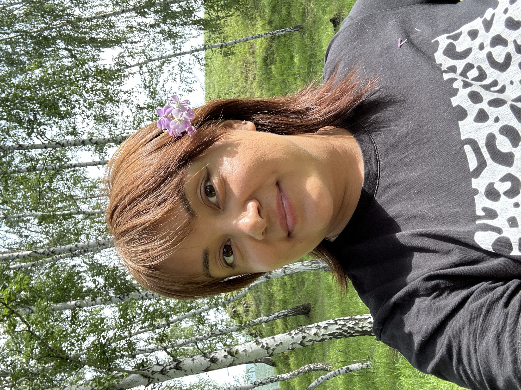 In an outdoor, natural setting with a forest of birch trees and a grassy hillside, a woman with reddish-brown hair, adorned with a small cluster of purple flowers over her right ear, looks directly at the camera. She is wearing a black t-shirt with a white, abstract pattern that resembles leopard print on the lower portion of the garment. The lighting suggests it is daytime, and the overall atmosphere is casual and relaxed. The background consists of the dappled sunlight filtering through the leaves of the birch trees, creating a bright, slightly hazy effect.