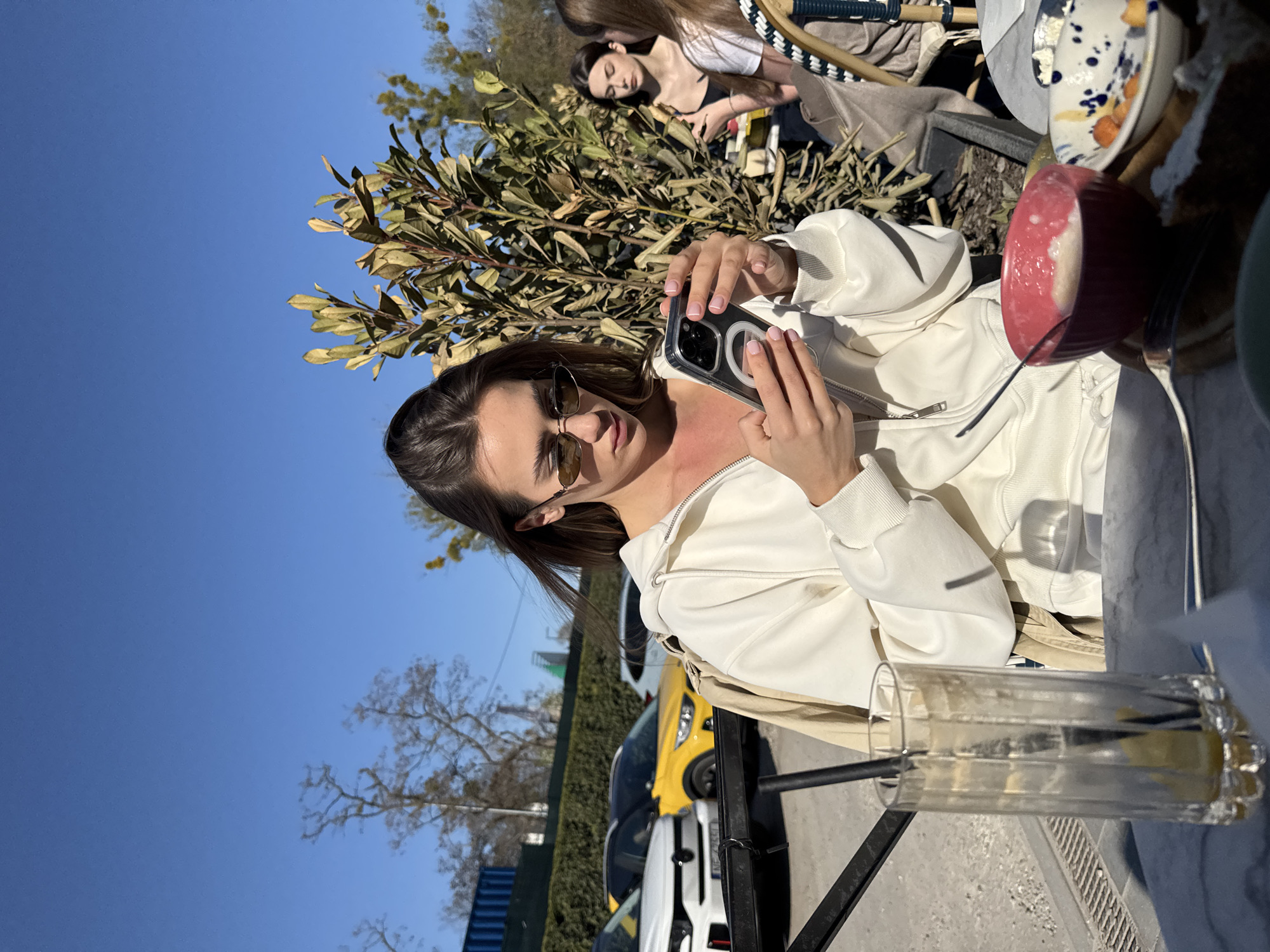 An outdoor, brightly lit scene captures a young woman at a cafe or restaurant patio, holding a smartphone with a magnetic ring on its clear case. She wears a white hooded sweatshirt and sunglasses, her brown hair pulled back. Her left hand, with light pink manicured nails, is visible on the phone. She appears to be looking towards the camera or at her phone.
Behind her, another young woman with dark hair sits at a table, partially obscured by a lush green bush. In the far background, a third person's head is visible. On the table in front of the main subject, there is a red bowl, patterned ceramic plates, a fork, and a tall clear glass containing a light-colored beverage with lemon slices and a dark straw.
The setting is an open-air area, possibly a sidewalk cafe, with a vibrant blue sky overhead, indicating a clear, sunny day. Trees are visible against the sky. In the background, beyond the patio, several cars are parked along a street, including a distinctive yellow sports car and a white car. The overall atmosphere is relaxed and casual, suggesting a daytime outing. There is no discernible text visible in the image. The location is contextualized as Warsaw, Poland.