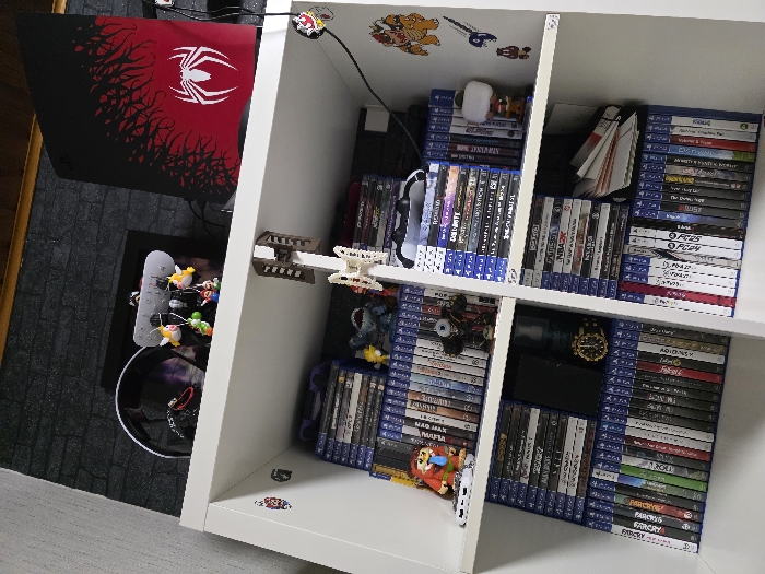 This image depicts an indoor scene within a residence in Zweibrücken, Germany, focusing on a white modular shelving unit positioned against a wall. The unit is primarily populated with gaming-related items.
The white shelving unit has at least three horizontal shelves and two vertical dividers, creating six visible cubbies and a top surface. The wall behind the unit features dark, possibly charcoal or black, faux-brick textured wallpaper or paneling. A dark wooden trim or ceiling is visible at the top edge of the frame.
On the top surface of the shelving unit, from left to right: a black over-ear gaming headset is present. To its right, a white display stand holds multiple small, colorful keychains, including characters resembling Mario and Luigi. Behind these, a black framed object is partially visible. On the far right, a black vertical gaming console, likely a PlayStation 5, stands upright. Its front panel is adorned with a custom red and black "Spider-Man" themed skin featuring a prominent white spider logo. Cables are visible connected to the console.
The cubbies of the shelving unit are organized as follows:
* The top-left cubby contains stacks of PlayStation 4 game cases, a small white electronic device, a figurine resembling Link from "The Legend of Zelda," and a dark, armored figurine.
* The top-right cubby holds stacks of PlayStation 4 game cases, with a white PlayStation DualShock 4 controller resting on top of one stack. A Funko Pop! figurine with a white shirt is positioned on the right.
* The middle-left cubby is filled with PlayStation 4 game cases. A dark-colored smartphone lies flat on top of some cases, next to a gold-colored wristwatch with a black bezel and a dark cylindrical object.
* The middle-right cubby contains more PlayStation 4 game cases, with a stack of horizontally oriented white-spined cases or books on the right.
* The bottom-left and bottom-right cubbies are filled with standing PlayStation 4 game cases.
Several stickers are affixed to the white shelving unit: a small red and white sticker on the upper left vertical divider, a red and white circular sticker near the top of the right vertical divider, and a collection of stickers on the far right side of the unit, including a large yellow and brown character resembling Bowser, a blue number "9", and smaller brown mushroom-shaped icons. Two white, multi-jointed plastic or 3D-printed objects are suspended from the underside of the top shelf, positioned centrally.