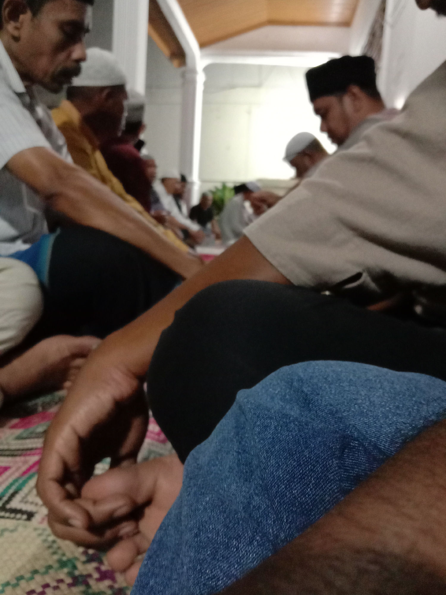 In a dimly lit interior, a group of men are seated on a mat, possibly in Lhokseumawe, Indonesia. Most wear white or black caps. One man wears blue jeans and another wears a blue shirt. The mat they are seated on has a patterned design. The background shows architectural columns and a light-colored ceiling. The men are oriented toward a central area, suggesting they may be engaged in a religious or social gathering.