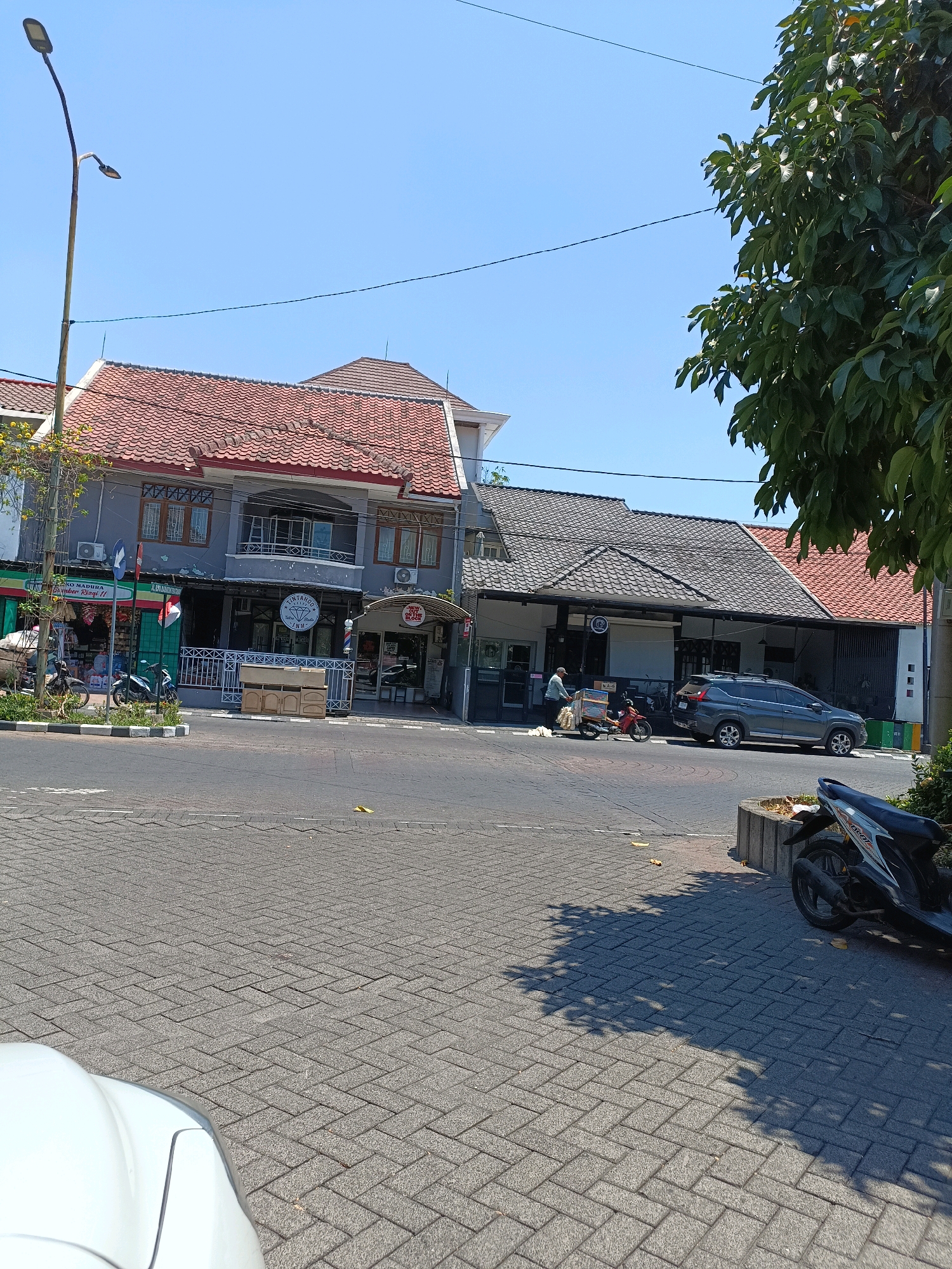 The image depicts a street scene in Surabaya, Indonesia, on a clear day. In the foreground, a paved road with intricate brickwork is visible, with the edge of a white car on the left. Further ahead, several buildings line the street. The building on the left has a gray facade with red-tiled roofs and multiple windows. A street lamp stands tall on the left side of the image.
To the right of the gray building, there is a lower, dark-tiled roof structure. A dark gray SUV is parked in front of this structure. A person is pushing a cart alongside a red motorcycle. In the distance, more buildings with red-tiled roofs are present. A large tree with dense green foliage is seen on the far right, casting shadows on the pavement. A parked motorcycle with a white and black design is in the lower right corner. Various signs and shop fronts are visible on the buildings, suggesting a commercial area. The sky is a clear, bright blue.