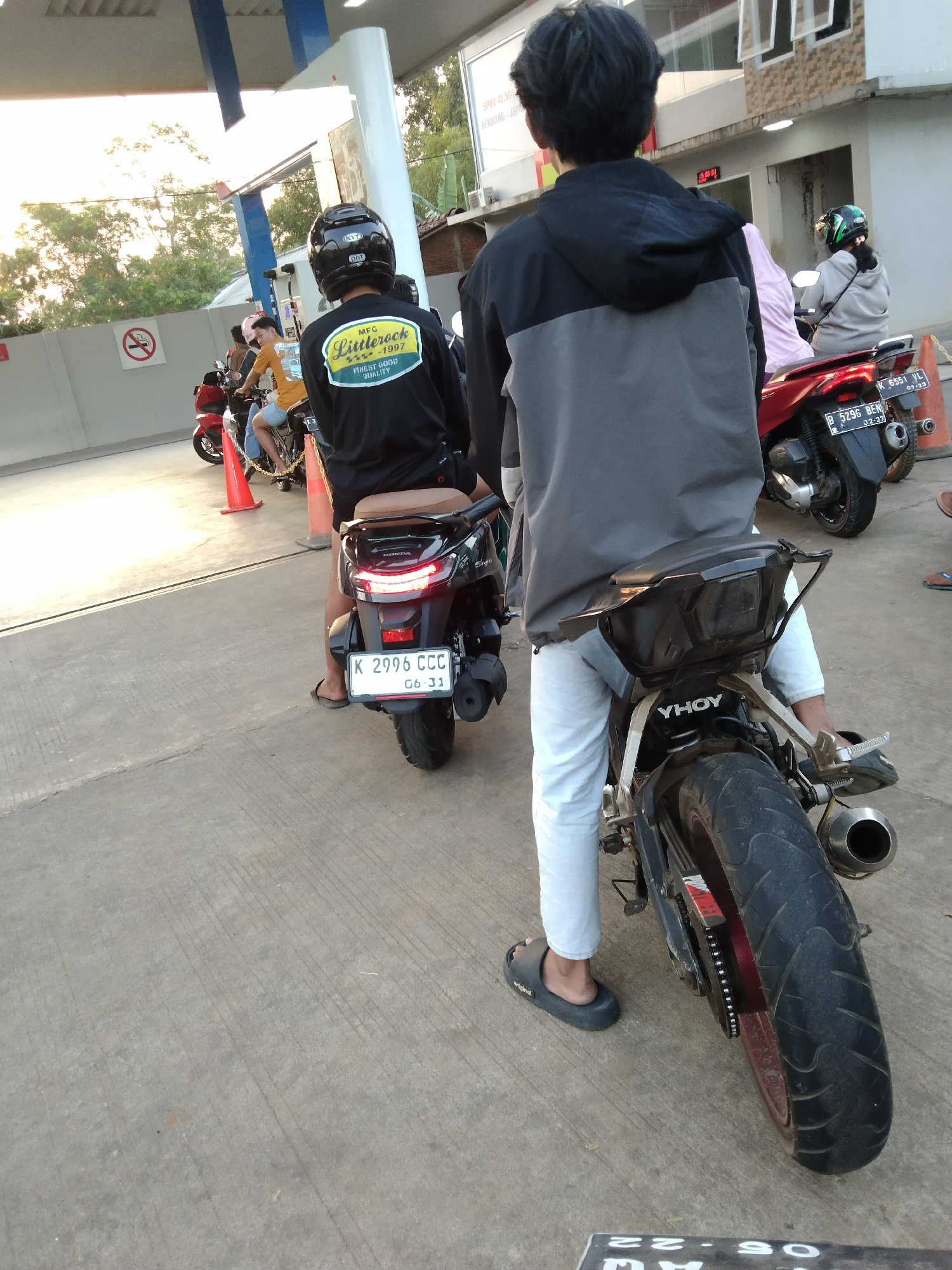 The image shows a street scene in Kembang, Indonesia, likely a gas station. In the foreground, a person wearing white pants and black flip-flops is seated on a black motorcycle, facing away from the camera. The motorcycle has a black seat and a license plate with the inscription "K 2996 CCC 06-31". Behind this motorcycle, another person is seated on a red motorcycle, also facing away from the camera. This person wears a black shirt with a yellow and green circular logo that reads "MFG LittleRock FINEST GOOD QUALITY -1997". The motorcycle has a license plate that is partially obscured. In the background, other motorcycles and people are visible, as well as a building and trees. The lighting suggests it is daytime, possibly late afternoon due to the warm glow.