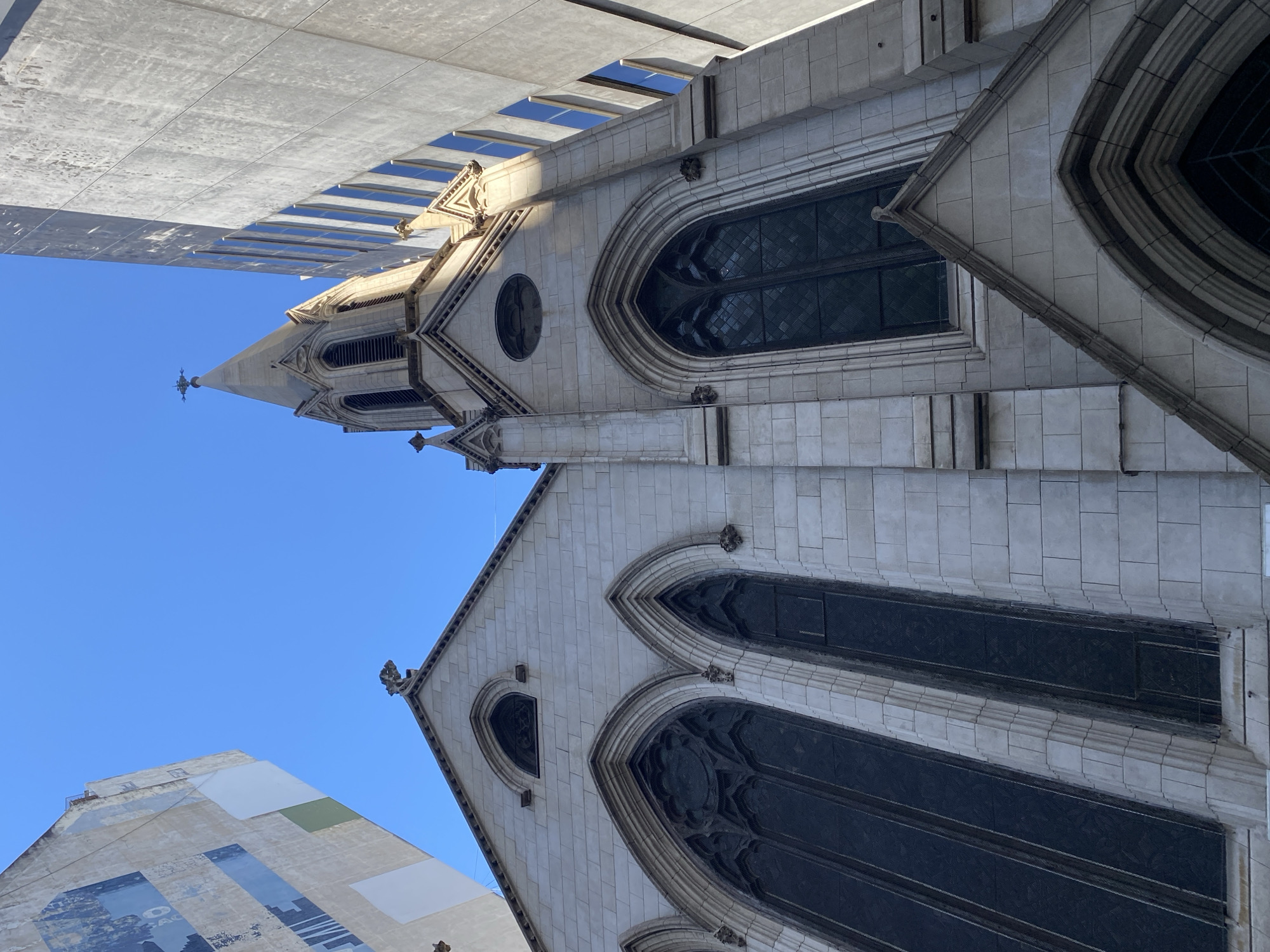 The image displays a majestic stone church, predominantly light-colored, seen from a low-angle perspective looking upwards. The photograph is oriented vertically, showing the church building reaching towards a clear blue sky.
The central subject is the ornate church, featuring a tall, slender spire topped with a finial, likely a cross. The architecture suggests a Gothic Revival style, characterized by intricate stonework, pointed arches, and decorative elements. Numerous arched windows, filled with dark, possibly stained or gridded glass, punctuate the facade. A circular rose window is visible on the tower section. Detailed carvings and smaller sculptural figures, resembling gargoyles, adorn various points of the building, particularly around the windows and cornices. The stone blocks forming the church's walls are meticulously laid, exhibiting texture and a weathered appearance.
Contrasting with the historic church are modern buildings visible in the background and foreground. To the upper left, a large, concrete-clad high-rise with horizontal window slits casts shadows onto itself. To the lower left, another contemporary structure features a facade covered in large, patchy panels, some with faint, dark graphics or text, including what appears to be "AG" and a cityscape silhouette.
The scene is bathed in bright daylight, indicated by the vibrant blue sky and distinct shadows cast on the church's surfaces, suggesting a sunny day, likely mid-morning or mid-afternoon. There are no people visible, and no specific activity or event is taking place, focusing solely on the architectural details and the interplay of different building styles against the sky. The location context for this architectural composition is Buenos Aires, Argentina.