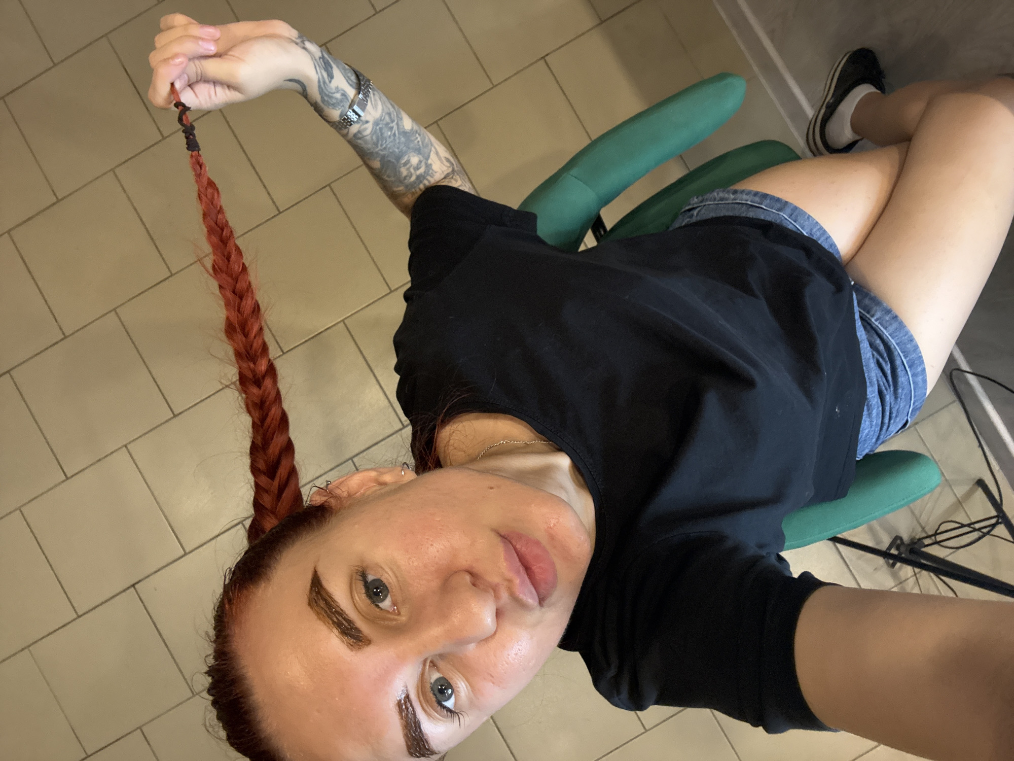 The image shows a woman with red braided hair sitting in a green chair indoors. She is wearing a black t-shirt and denim shorts. Her left arm has tattoos. Her right hand is holding the end of her braid. The background appears to be a tiled floor. The image was taken in Rostov-on-Don, Russia.