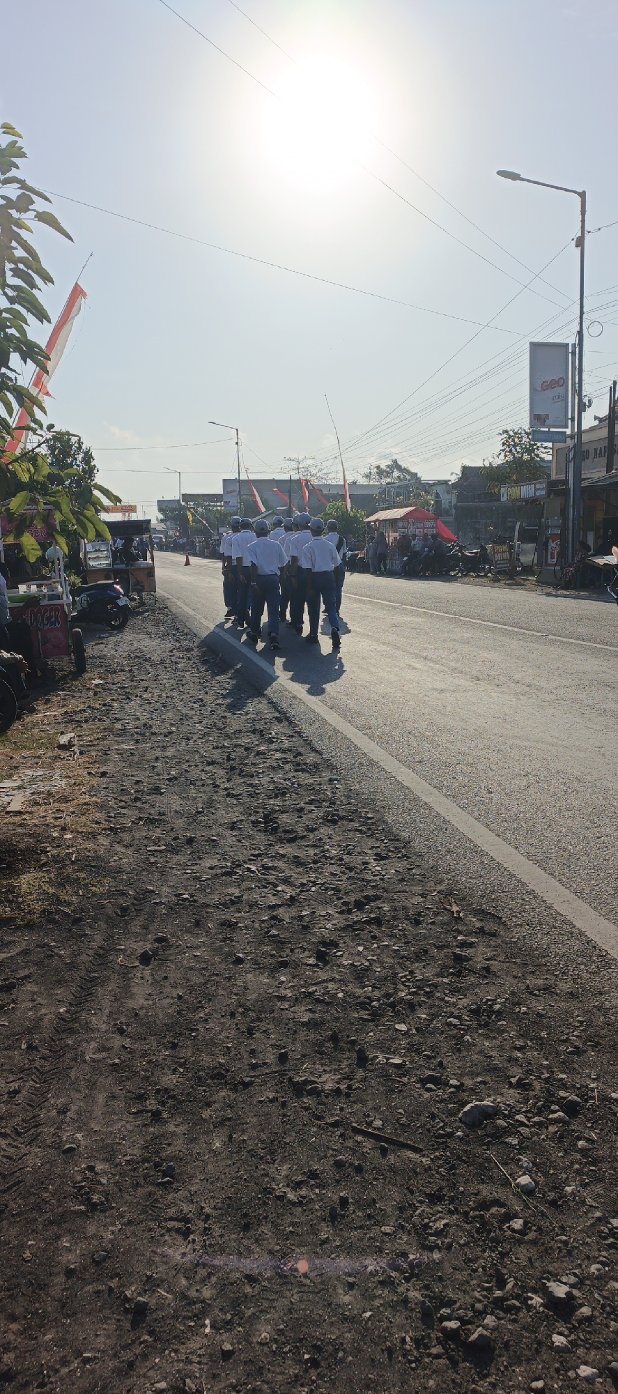 In Kasiyan, Indonesia, on a paved road under a bright, sunny sky, a group of approximately ten young men in white shirts and dark pants walk in formation. They are dressed in what appears to be a school uniform, with some wearing caps. To the left of the road, there is a narrow, unpaved area with loose gravel and dirt. In this area, several food carts and a parked motorcycle are visible. Red and white Indonesian flags are displayed on poles along the roadside, suggesting a patriotic or festive occasion. In the background, buildings and utility poles line the street. The overall scene depicts a procession or organized activity taking place outdoors.
