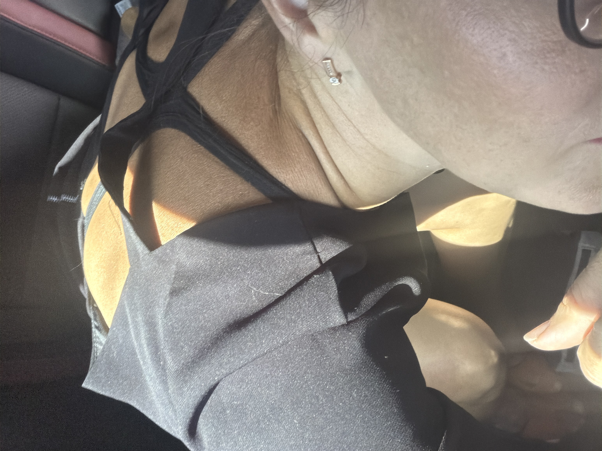 The image shows a close-up, indoor shot of a person in what appears to be a car. The person is wearing a black top with thin straps. Sunlight is casting shadows across the scene, highlighting the texture of the skin and clothing. A small silver piercing with a stone is visible on the person's neck. The background shows the dark interior of a car, with hints of red trim on a seat. There is no information in the image to determine the location in Votkinsk, Russia.