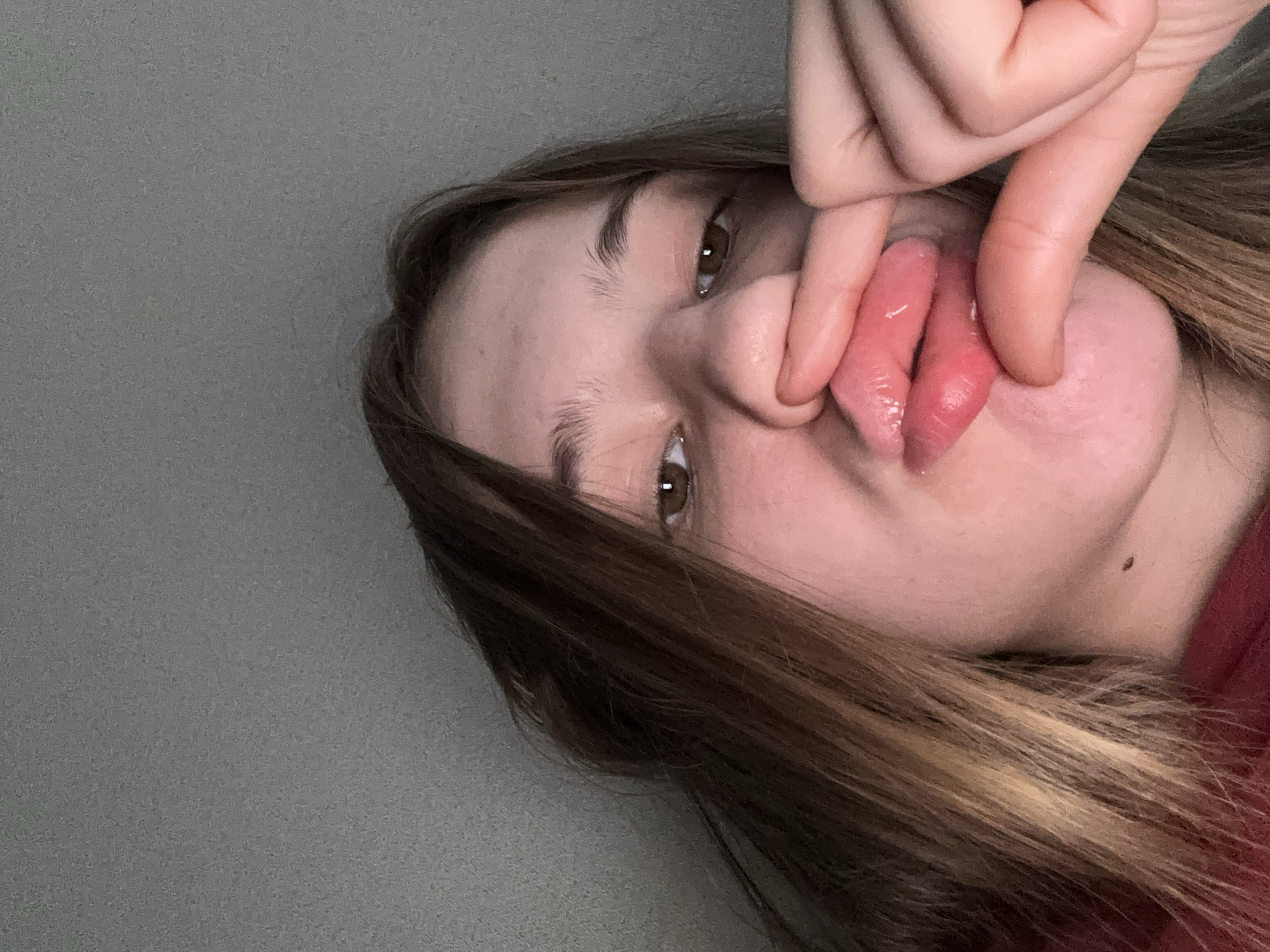 The image shows a close-up, low-angle, indoor selfie of a young person with light brown hair. Their face is partially obscured by their hand, which is pulling down their lower lip. The background is a plain, textured gray wall. The person is wearing a red garment. There are no discernable elements in the image that would allow for the localization of the scene to Vsevolozhsk, Russia.
