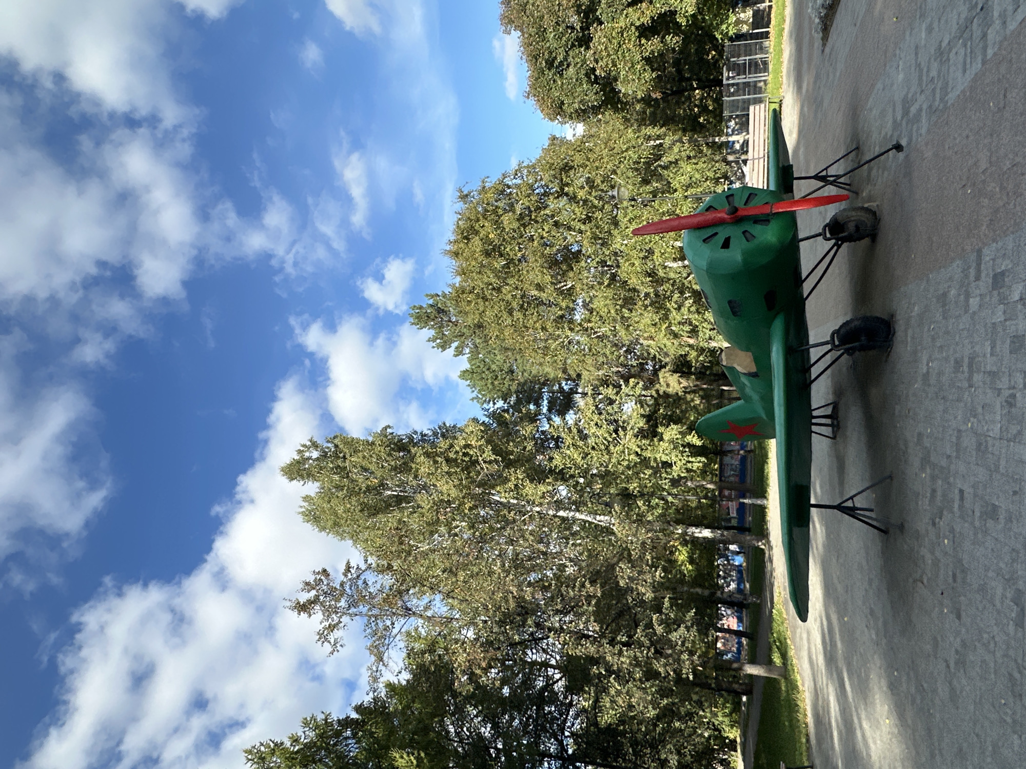 An outdoor, eye-level, full shot captures a small, bright green, vintage-style airplane sculpture on a paved area. The airplane has a red propeller and a red star emblem on its tail. It is propped up on thin black legs with wheels. Behind the sculpture is a line of trees with green and yellow foliage, suggesting it is autumn. The sky above is blue with scattered white clouds. The paved area in front of the plane has a brick-like pattern. To the left of the airplane, a bench is visible. Further back, behind the trees, there appears to be a fence and some indistinct structures, possibly buildings or playground equipment. The lighting suggests it is daytime.