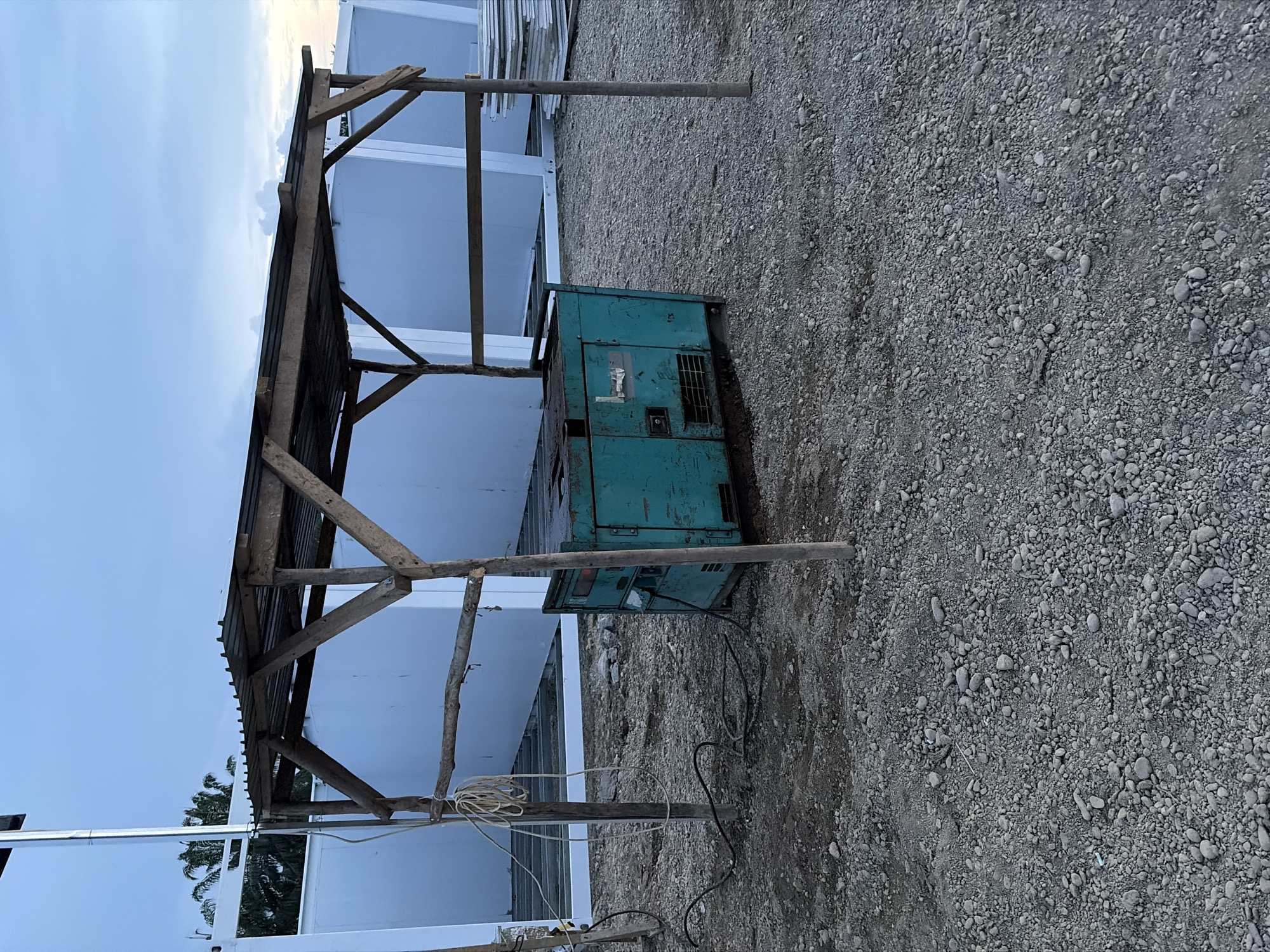 The scene depicts a gravel lot in Simpangempat, Indonesia. A weathered, teal-colored generator is the central object, situated near a makeshift wooden structure providing rudimentary shelter. The structure's supports are rough-hewn posts, and the roof appears to be corrugated material. Two electrical cables are plugged into the generator and extend along the ground. In the background are white, rectangular buildings of indeterminate purpose. The sky is visible in the upper portion of the frame.