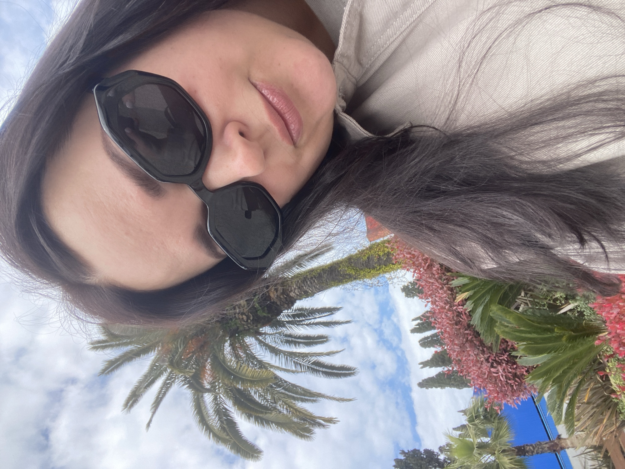 A selfie-style photograph presents a person, likely female, oriented along the left side of the frame. Her head is tilted, appearing to rest on her right shoulder or a surface, facing towards the top of the image. She wears dark, stylish sunglasses that obscure her eyes, and her lips are subtly colored with light pink lipstick. Her long, dark hair flows downwards and across the lower part of the frame, resting on a light-colored, possibly beige or grey, textured jacket or shirt. Her expression appears relaxed.
The background, visible on the right half of the image, is an outdoor scene under a bright, partly cloudy sky, characteristic of Sochi, Russia. A tall palm tree stands prominently, its trunk extending vertically from the mid-right to the upper-right corner, adorned with green foliage and some climbing vines. Its fronds spread out towards the lower right. Below and to the right of the palm tree, vibrant red-pink and green tropical foliage creates a colorful display. A segment of a bright blue structure or wall is visible at the very bottom right corner, peeking from behind the plants. The sky, with scattered white clouds against a clear blue, is primarily seen in the upper right section of the background. The time of day is daytime. No text is visible.