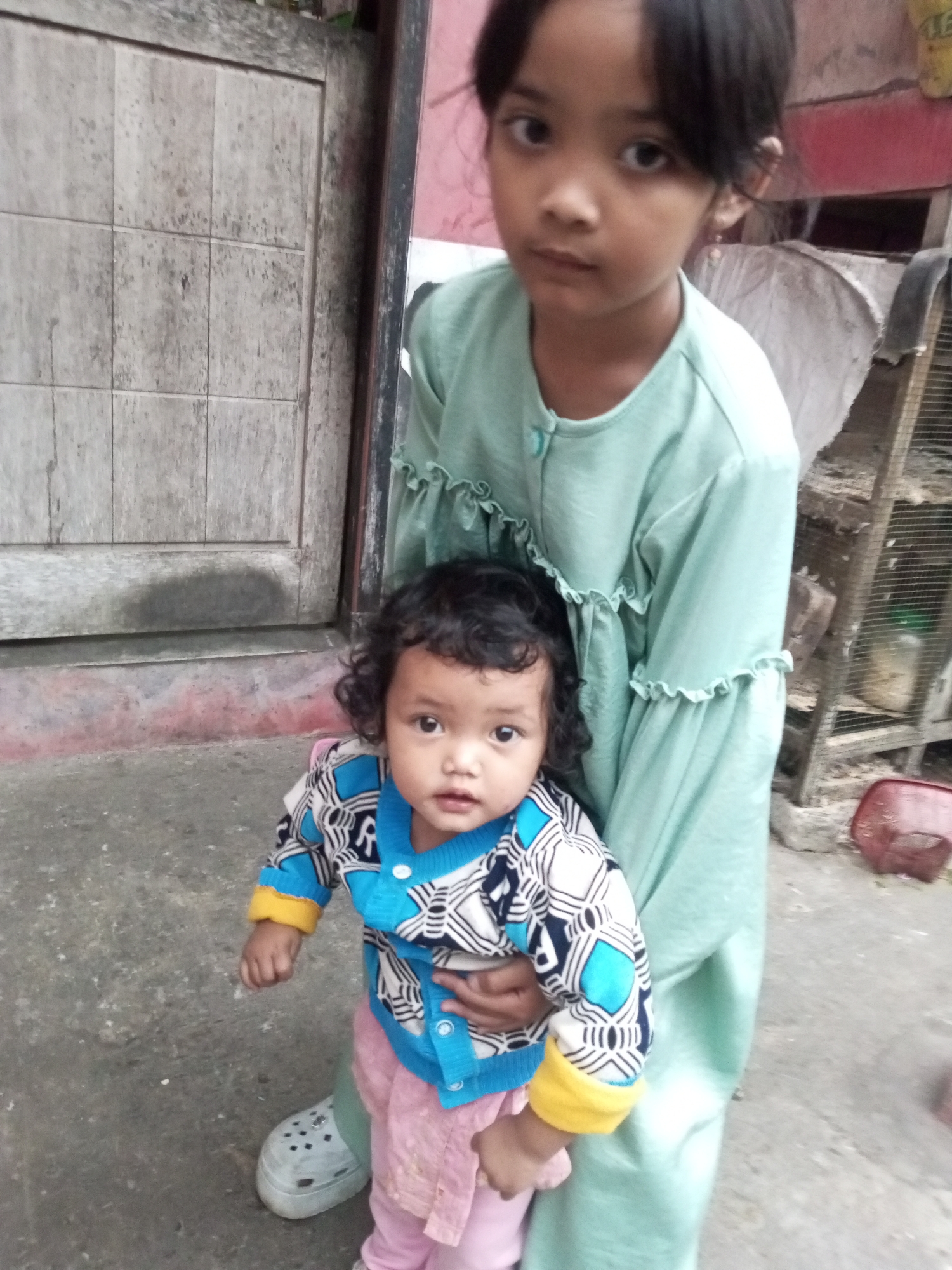 In Andongsili, Indonesia, a young girl in a mint green dress stands, holding a toddler wearing a blue and white patterned sweater and pink pants. The toddler has curly dark hair and a round face. They are positioned in front of a weathered wooden door with a tiled pattern. The ground is concrete, and to the right, a birdcage is visible.