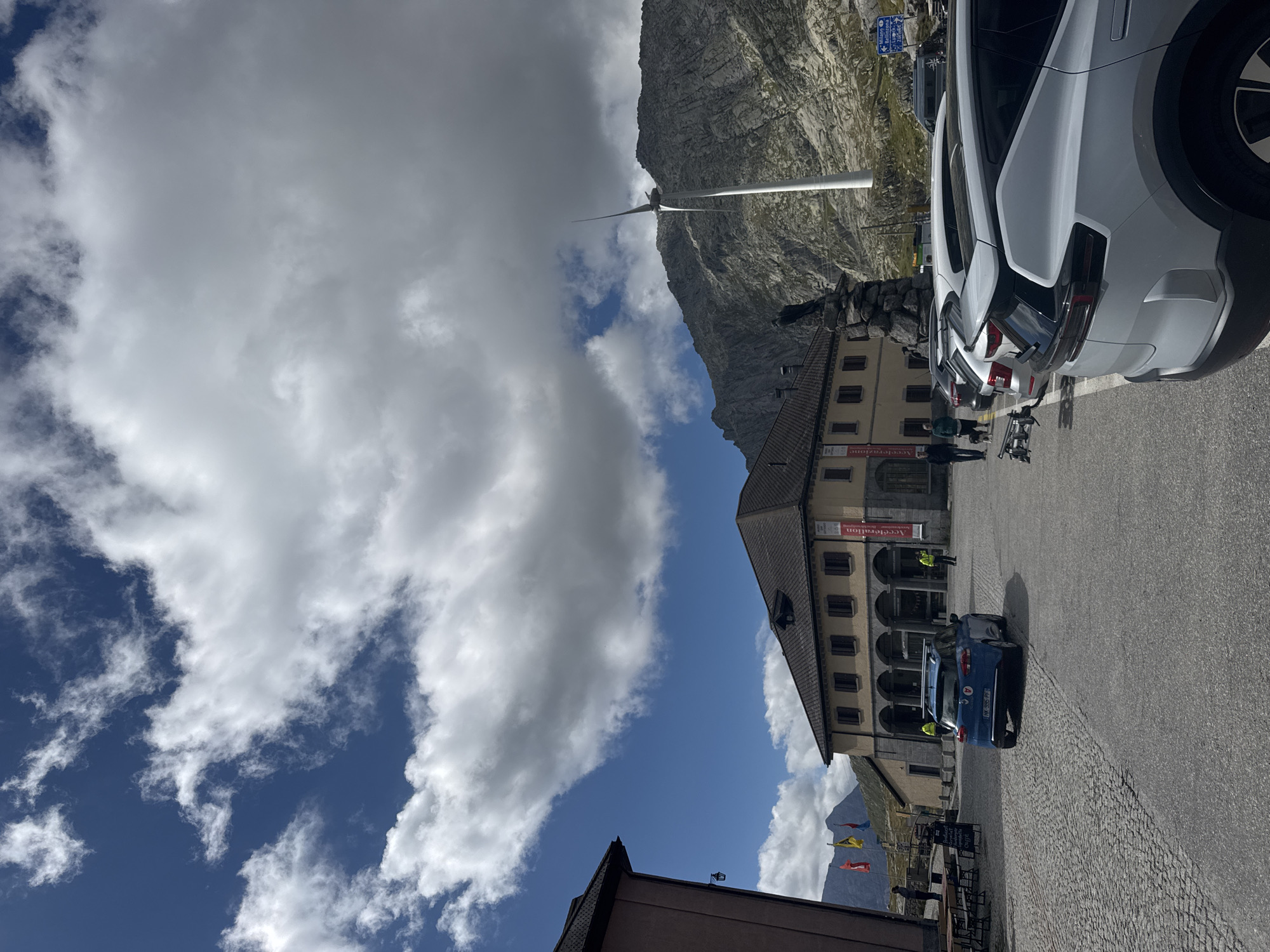 The image shows a high-altitude location with a building and parked cars in the foreground. A blue car is parked in the middle of a cobblestone area, facing away from the viewer. Several white cars are parked in a lot to the right. Behind the cars and building, a steep, rocky mountain range dominates the background. A large wind turbine stands on the mountainside, to the right of the central building. The sky is a mix of blue with scattered white and gray clouds. A statue of a bird is visible on a stone base near the building. The building appears to be a visitor center or a restaurant, with arched doorways and windows. There are a few people visible, some standing near the building and others near the parked cars. The scene suggests a scenic viewpoint or a rest stop in a mountainous region. The location is likely the Gotthard Pass, given the context of Airolo, Switzerland, as it is a significant mountain pass with a visitor center and is known for its dramatic landscapes and wind turbines.