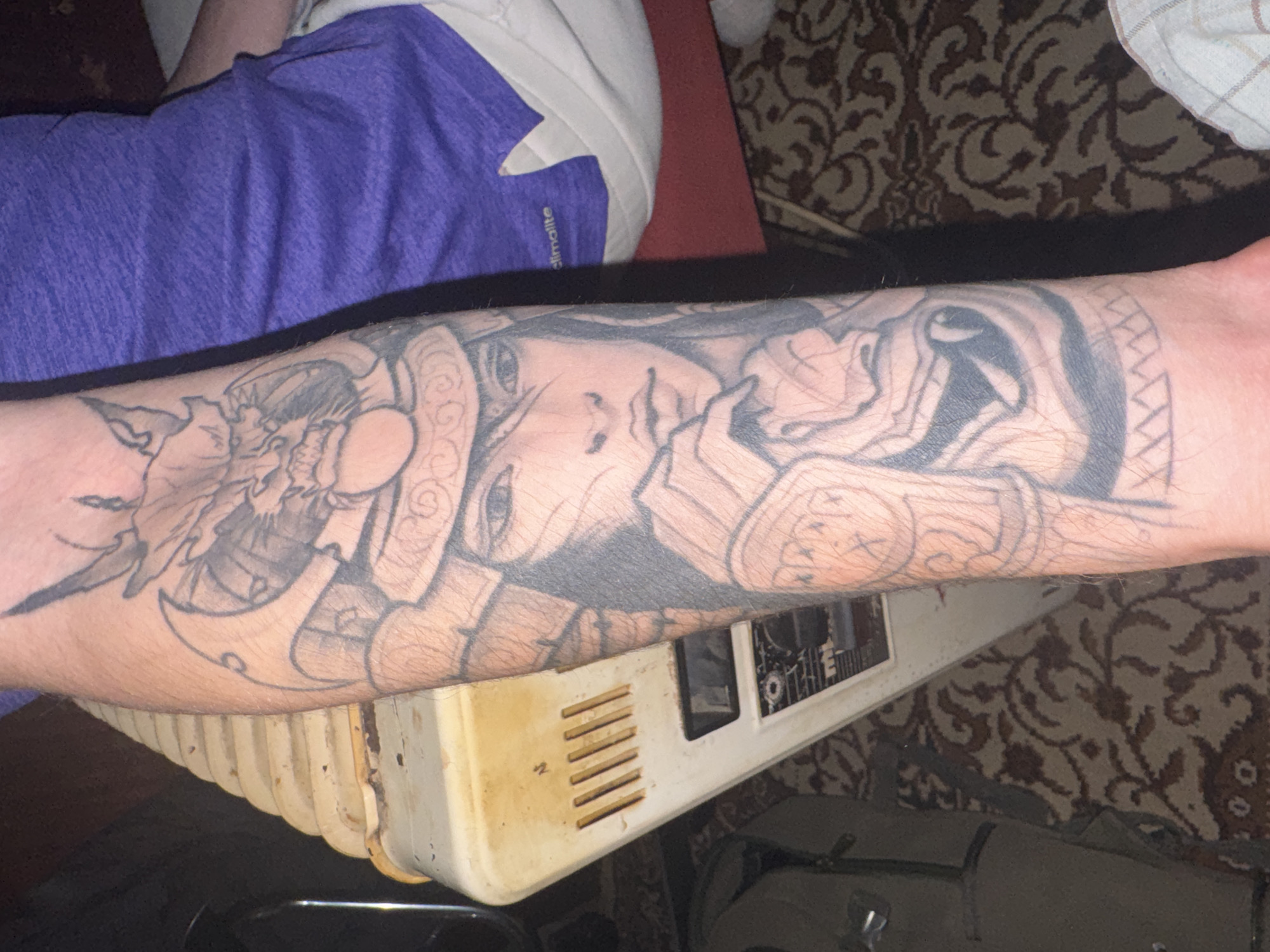 An arm with a tattoo is resting on top of a dirty, white, box-shaped object. The tattoo appears to depict a person's face and other complex designs. A portion of a person is visible in the upper left, wearing a purple shirt. The background includes a patterned rug and other indistinct objects. The location in the city of Unknown, Unknown is not discernible from the image.
