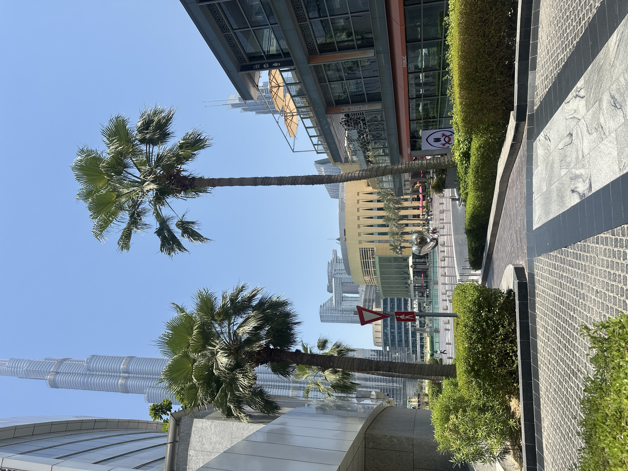 This is a ground-level, outdoor, eye-level shot of a public area in Dubai, United Arab Emirates, featuring modern architecture and landscaping under a clear blue sky. In the foreground, there are neatly trimmed green bushes and paved pathways made of stone or concrete. Two tall palm trees stand prominently in the middle ground.
Behind the palm trees, there is a body of water, possibly a fountain or canal, with people walking and interacting in the distance. Several modern buildings surround the water, with one large, yellow-gold building featuring a distinctive arched design. To the right, a multi-story building with balconies and an outdoor seating area is visible, with awnings providing shade. Signs with the word "PUBLIC" are displayed on this building.
In the background, the iconic Burj Khalifa skyscraper dominates the skyline, appearing as a tall, slender structure with a shimmering, glass facade. Other modern skyscrapers and buildings are also visible in the distance, contributing to the urban landscape of Dubai. A red and white triangular sign indicating a yield or warning is positioned next to a pedestrian crossing sign. On a banner hanging from a building, a cartoonish face with wide eyes and a red mouth is depicted. The overall scene conveys a sense of a bustling urban center with a blend of natural elements like palm trees and man-made structures.