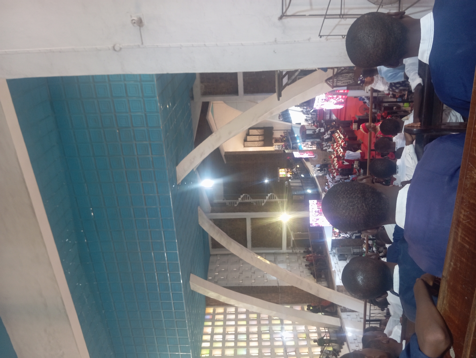 The image shows an interior view of a church, likely in Onitsha, Nigeria, judging by the appearance of the building and the people present. The foreground features the backs of several people, possibly children or teenagers, wearing blue and white uniforms seated in wooden pews. They appear to be part of a congregation or group observing an event taking place at the front of the church. In the background, the church interior is visible, filled with a large crowd of people. There is a stage or platform at the front, with individuals dressed in red and white robes, likely officiating a religious ceremony or event. Screens displaying video feeds are visible above the crowd. Architectural details of the church include a blue, tiled ceiling, white structural support beams, and colored glass windows providing some natural light. A spiral staircase can be seen at the back of the church. The overall atmosphere suggests a significant religious gathering or service.