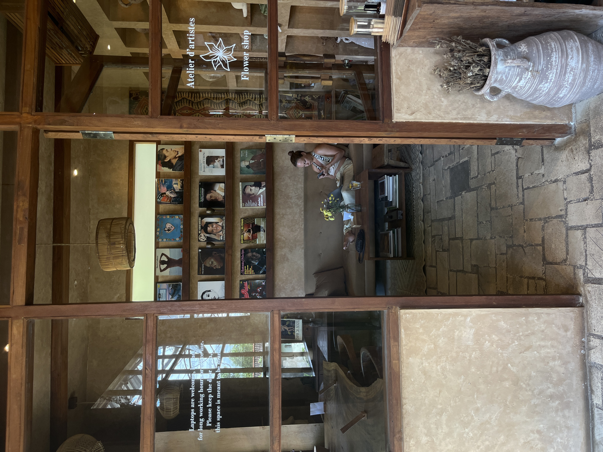 The image captures an interior view of "Atelier d'artistes Flower shop," a business in Kuta Selatan, Indonesia. The shop's entrance is framed by a wooden structure with glass panels. The glass reflects images of the shop's interior, including a woman seated with a phone, shelves displaying vinyl records, and a wooden table with yellow flowers. A woven lampshade hangs from the ceiling. The exterior includes a large, decorative vase filled with dried flowers. A sign on the glass states, "Laptops are welcome, for long working hours or..." and continues with a request to keep the door open for the shared space. The flooring outside the shop appears to be paved with stone tiles.